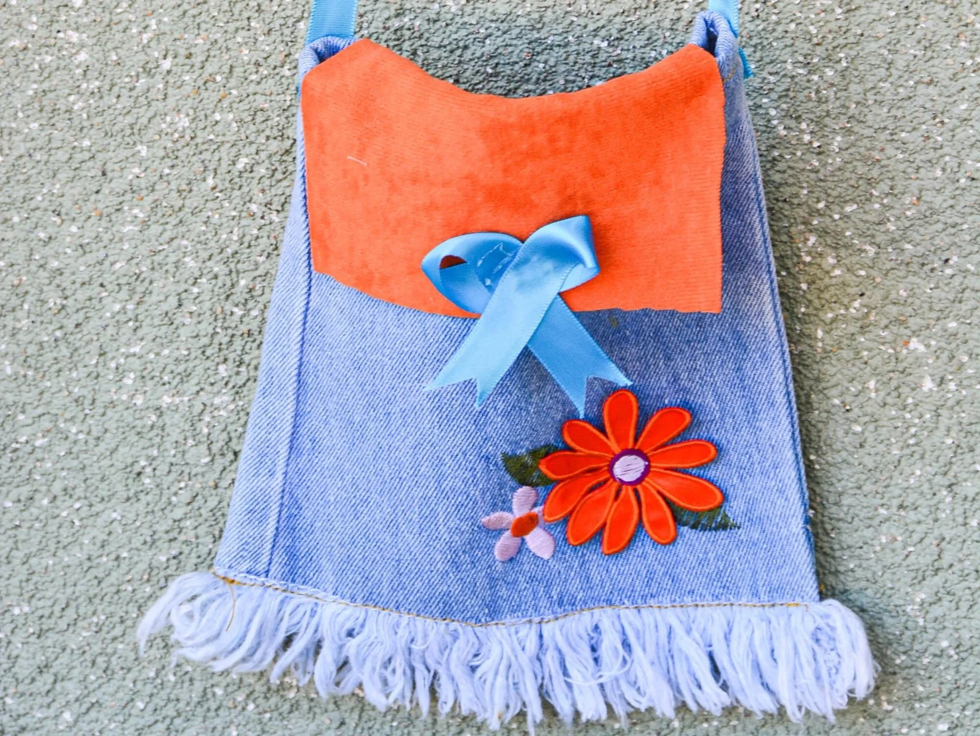 Decorative fabric item resembling a small bag or pouch with a blue ribbon, floral embroidery, an orange flower, and fringed bottom edge.
