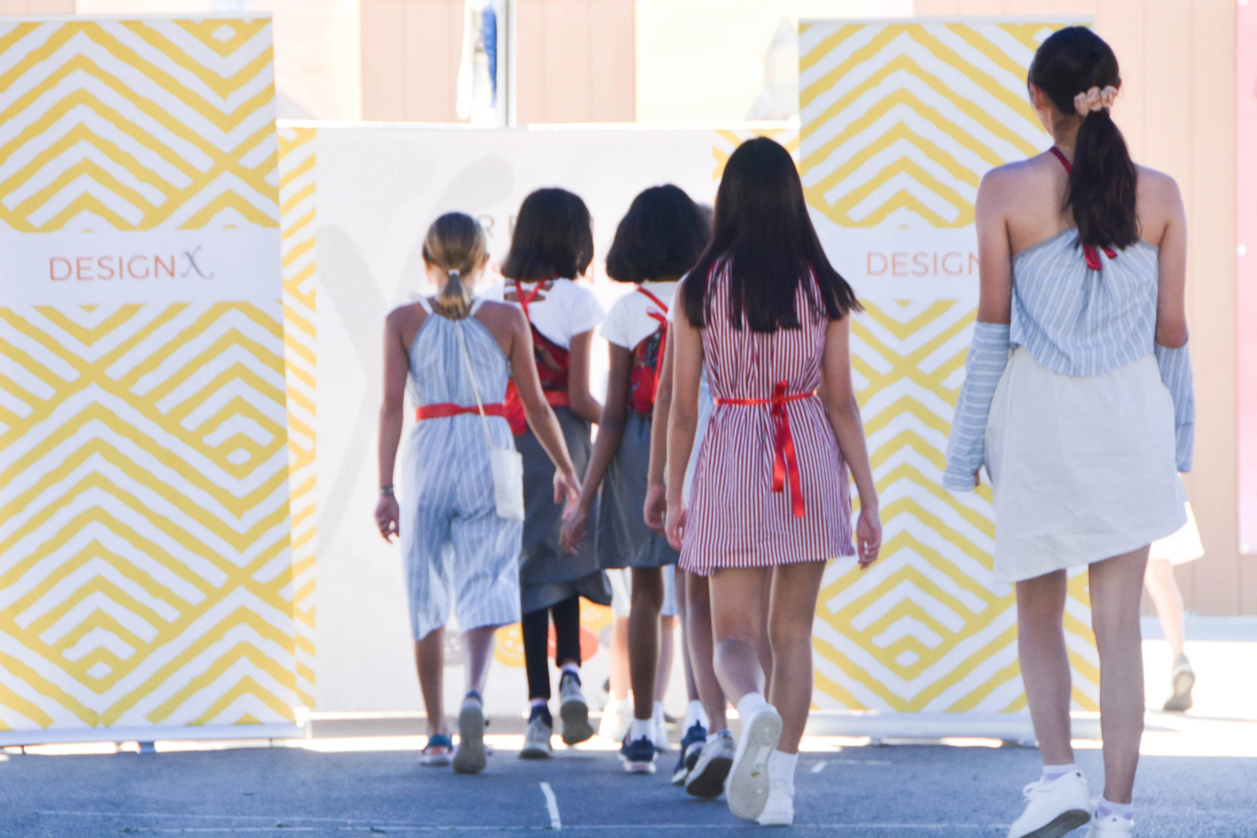 Group of children walking through an entrance at a design event, with a white and yellow patterned backdrop and the word 'DESIGNX' visible.