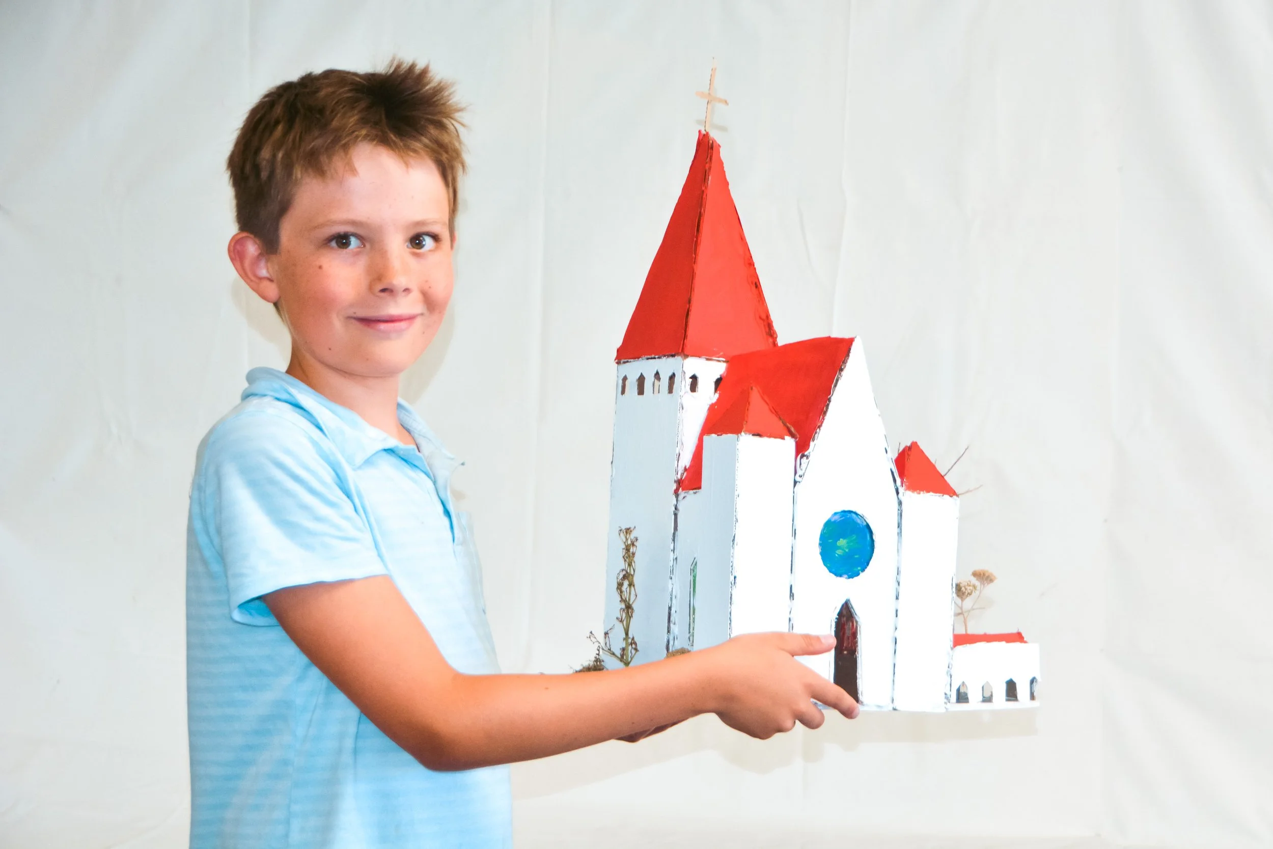 A young boy smiling and pointing at a small model of a church or castle with red roofs and a blue circular window, held in front of a plain white background.