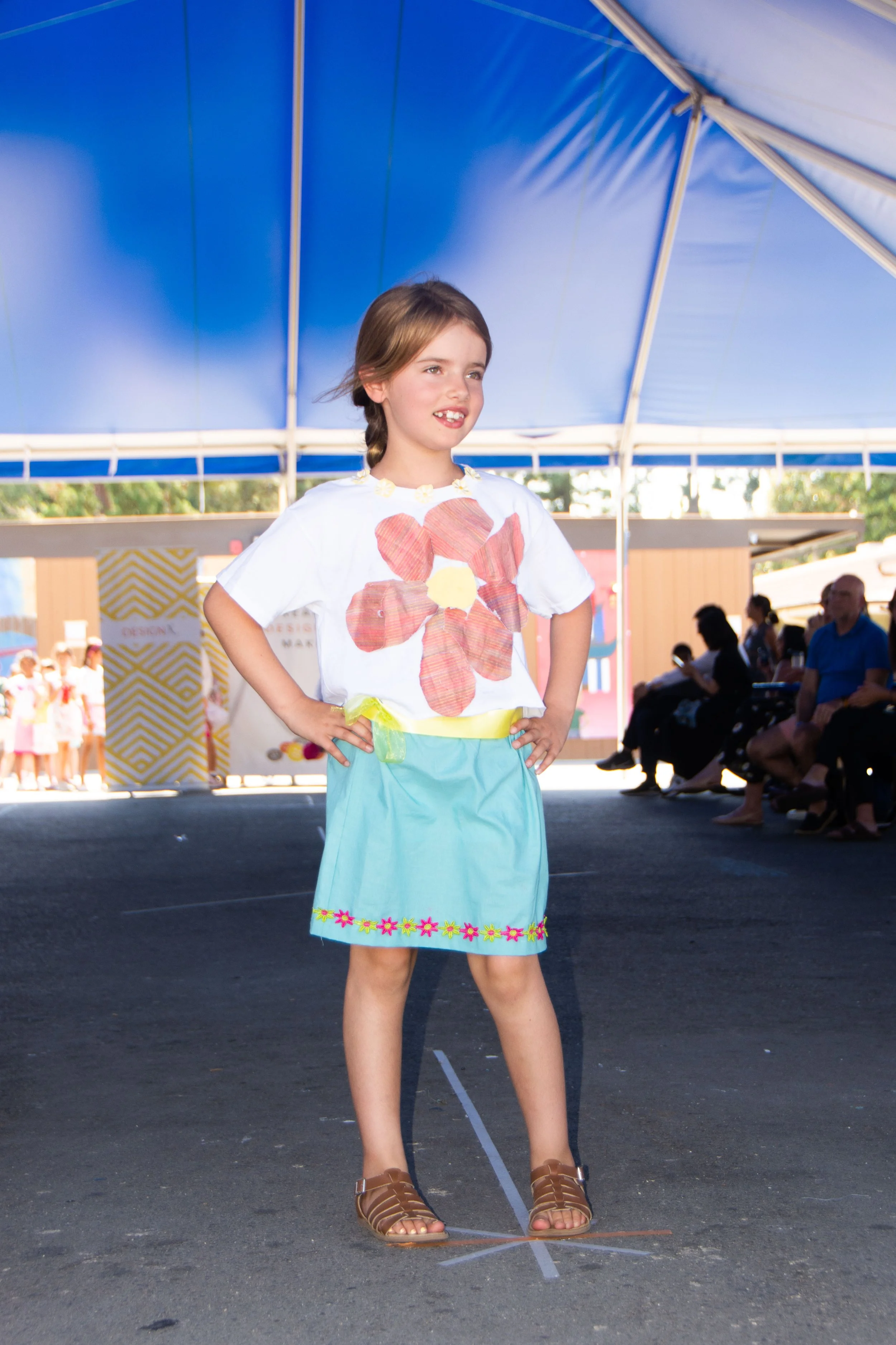 A young girl standing confidently with her hands on her hips during a fashion show, under a blue canopy, wearing a white T-shirt with a large flower print, a light blue skirt with floral embroidery, and brown sandals, with an audience seated nearby.