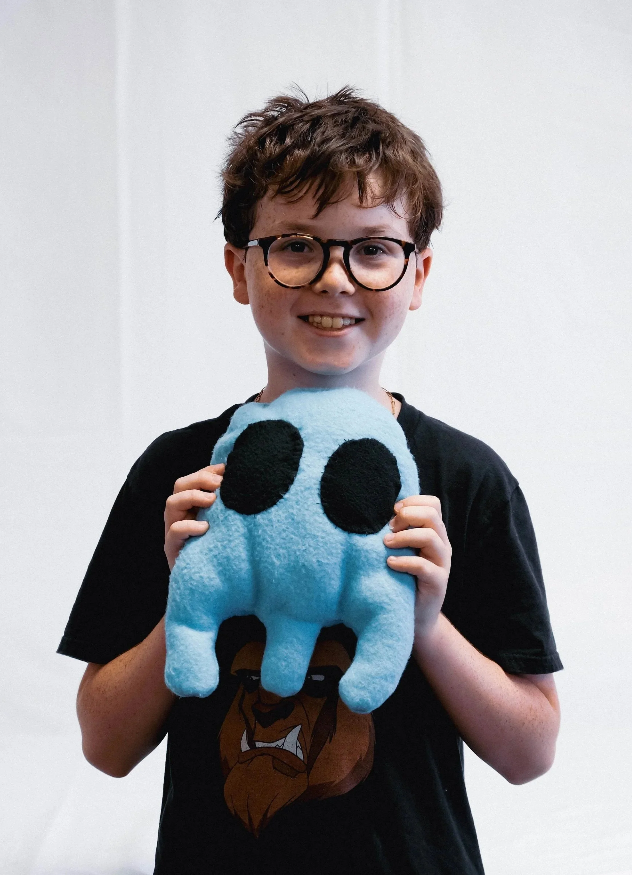 A young boy with glasses and short brown hair, smiling and holding a blue stuffed toy with black eyes and legs, against a plain white background.