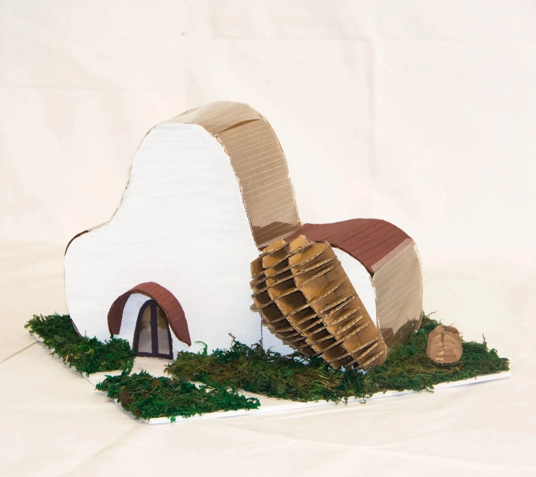 Model of a house made from cardboard with two rounded roofs, a door, and a garden area with moss and other natural materials.