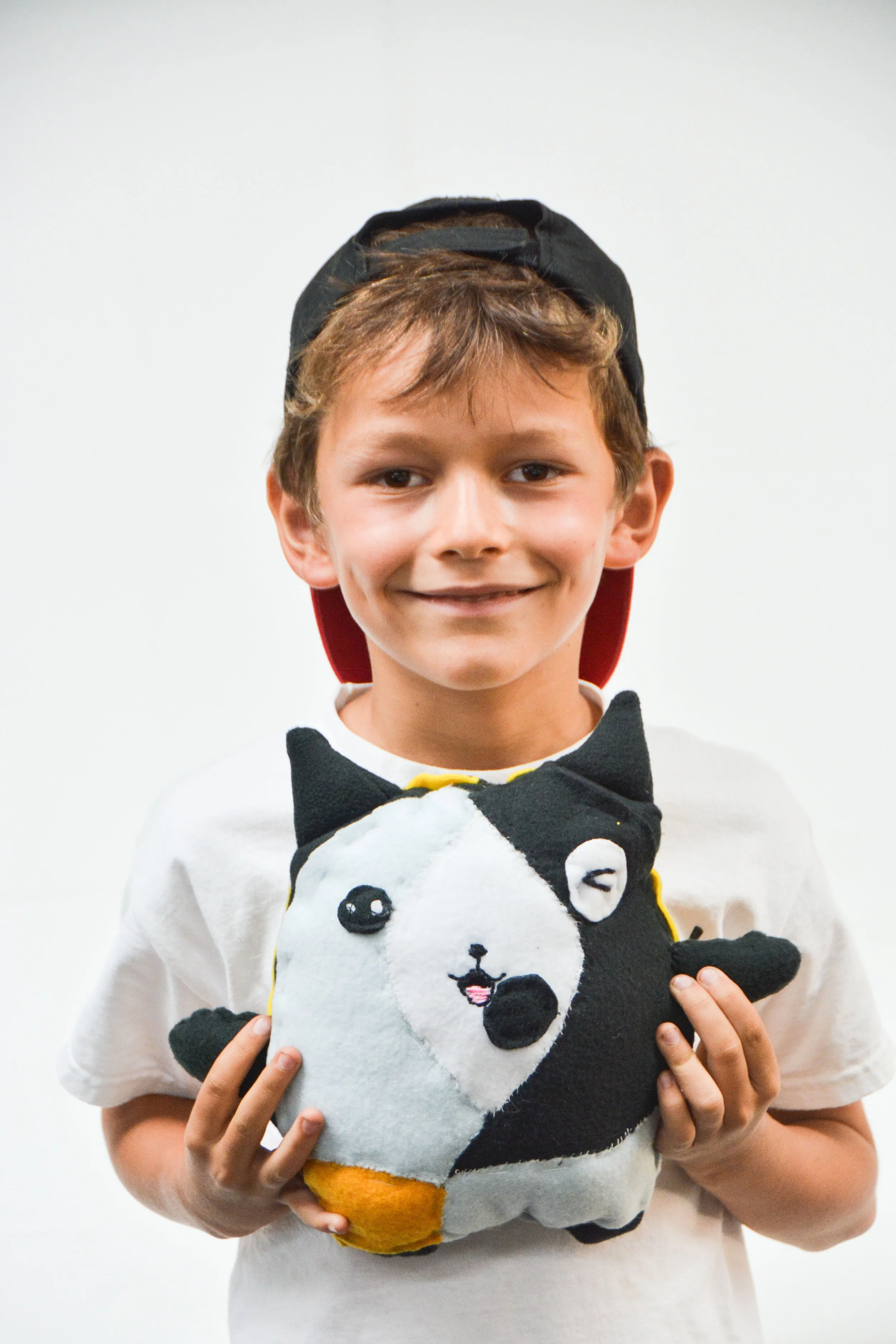 A smiling young boy wearing a black baseball cap backwards and a white t-shirt, holding a plush toy shark with a face resembling a cartoon dog, against a plain white background.