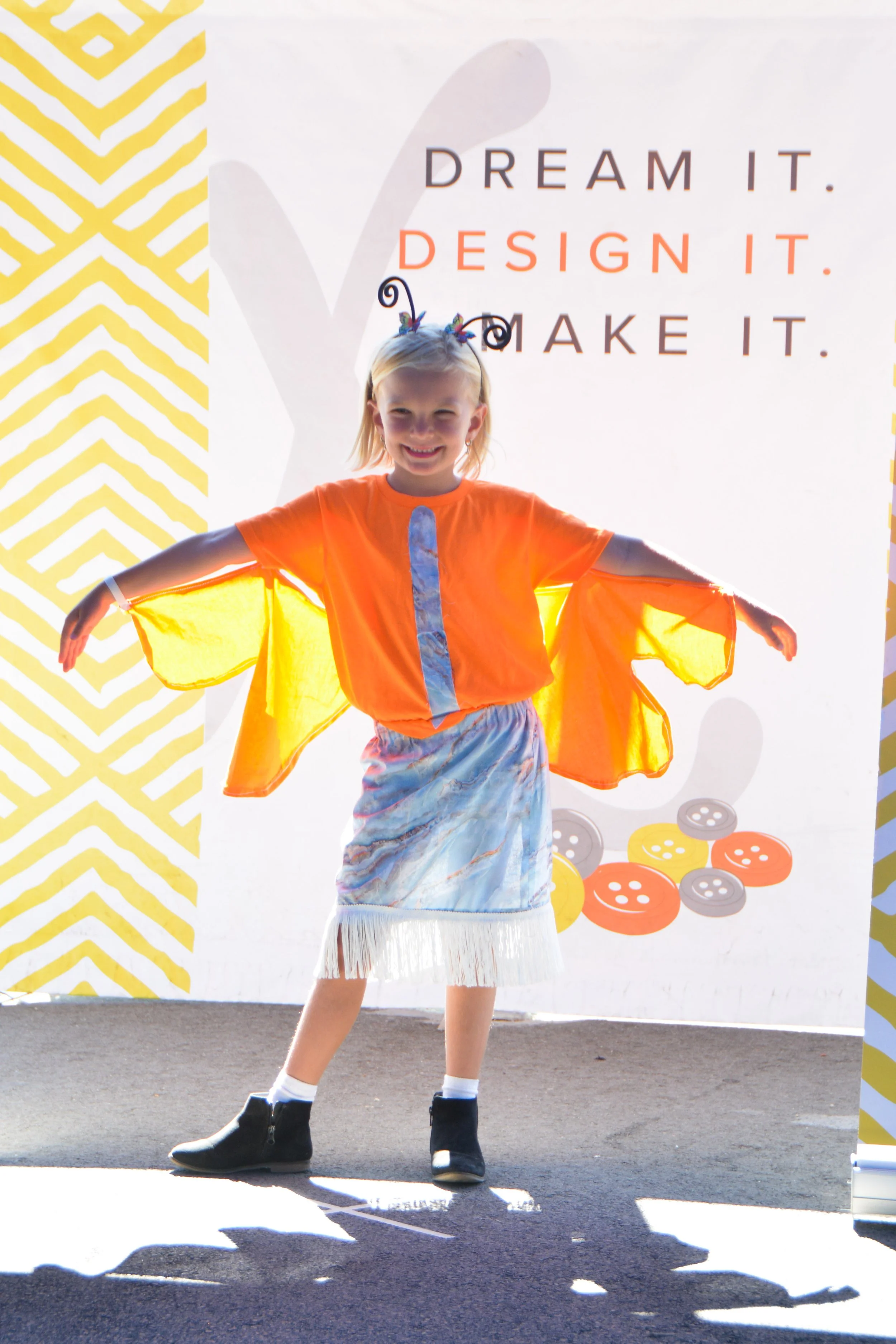 A young girl dressed in an orange top with blue and white skirt, standing with her arms outstretched in front of a backdrop with motivational text. The backdrop says, 'Dream it. Design it. Make it.' and features colorful buttons.