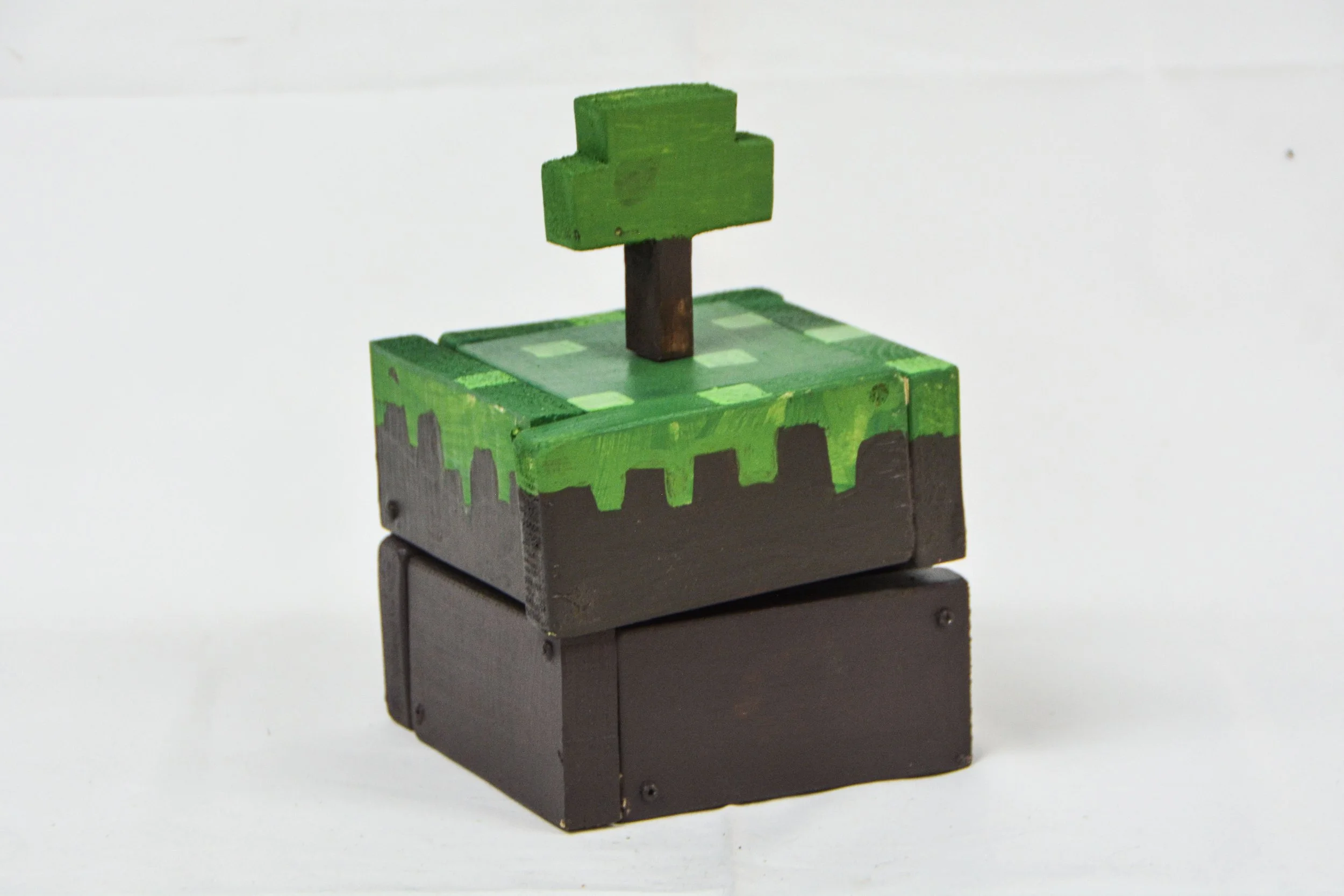 A small wooden block sculpture of the Minecraft grass block with a tree on top, painted in green, black, and brown, set against a plain white background.