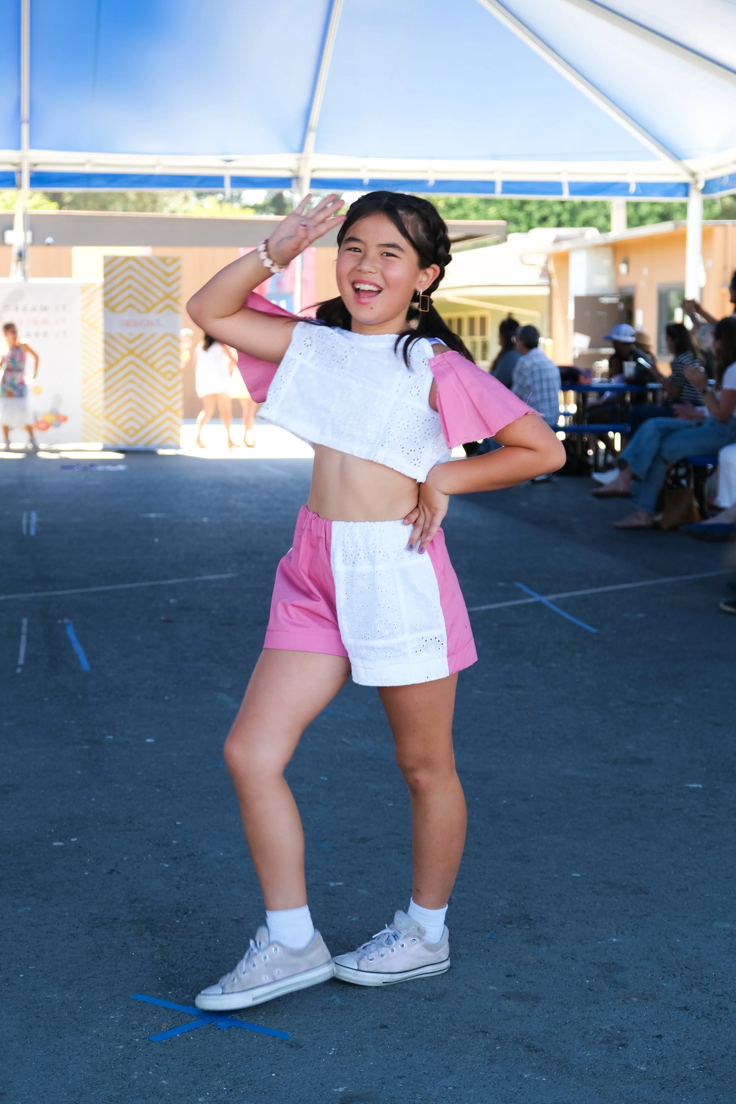 A young girl in pink shorts and a white top with pink sleeves, smiling and striking a pose during an outdoor event under a blue and white canopy, with people seated in the background.