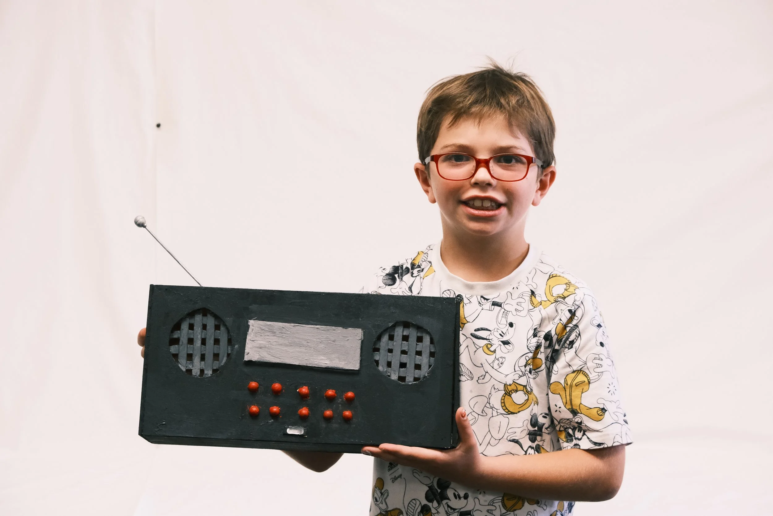 A young boy with glasses and a Mickey Mouse shirt holding a black painted cardboard radio with red buttons and a silver rectangle on the front, in front of a plain white background.