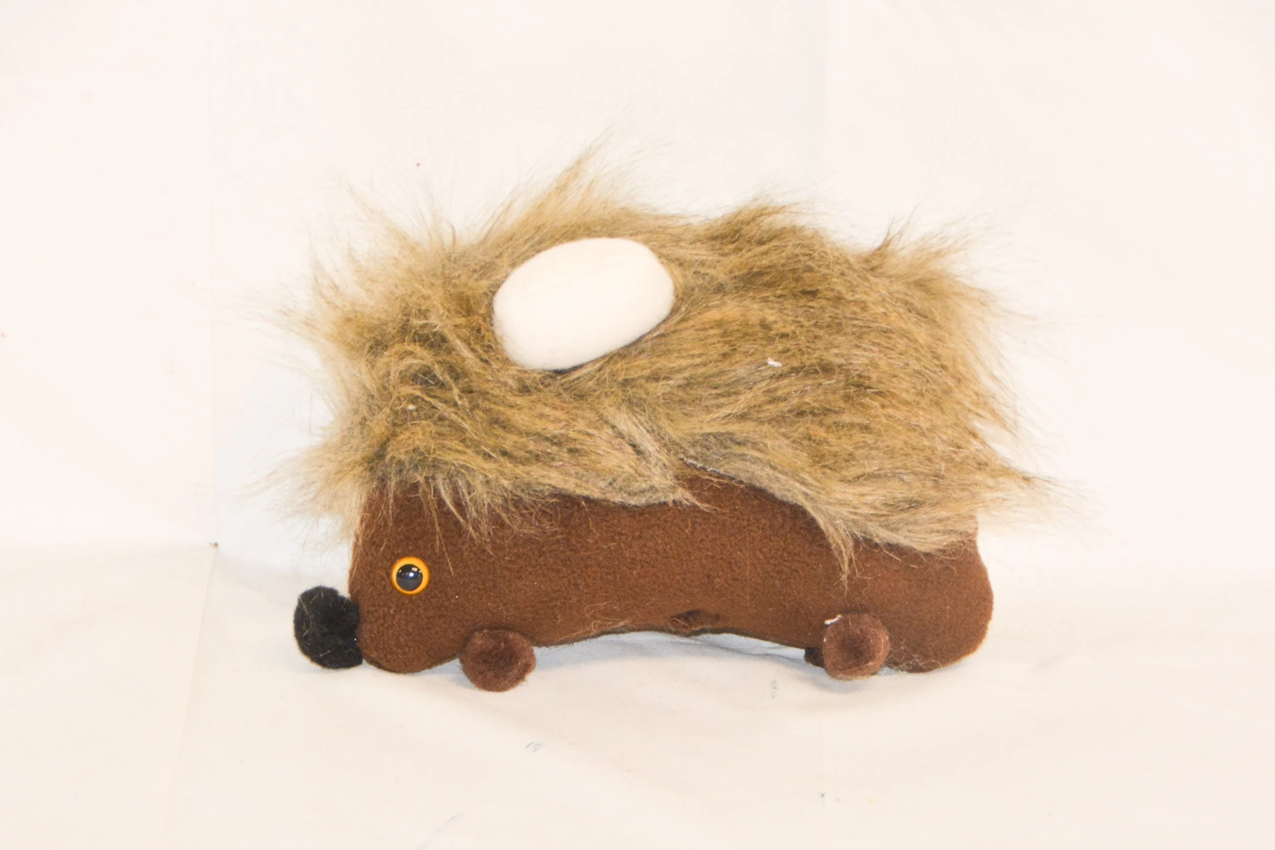A plush toy designed to look like a hedgehog with fur on top, a small black nose, a googly eye, and holding an oval-shaped object on its back, set against a plain white background.