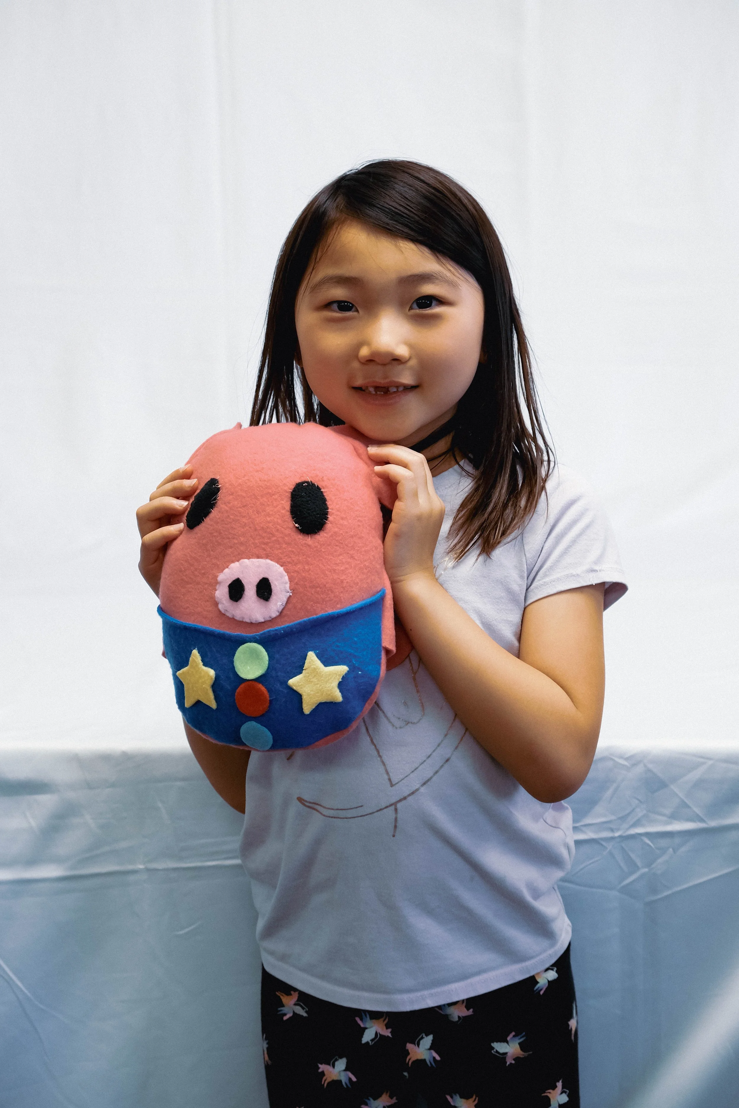 A young girl holding a pink plush pig wearing a colorful star and circle decorated outfit, smiling at the camera.