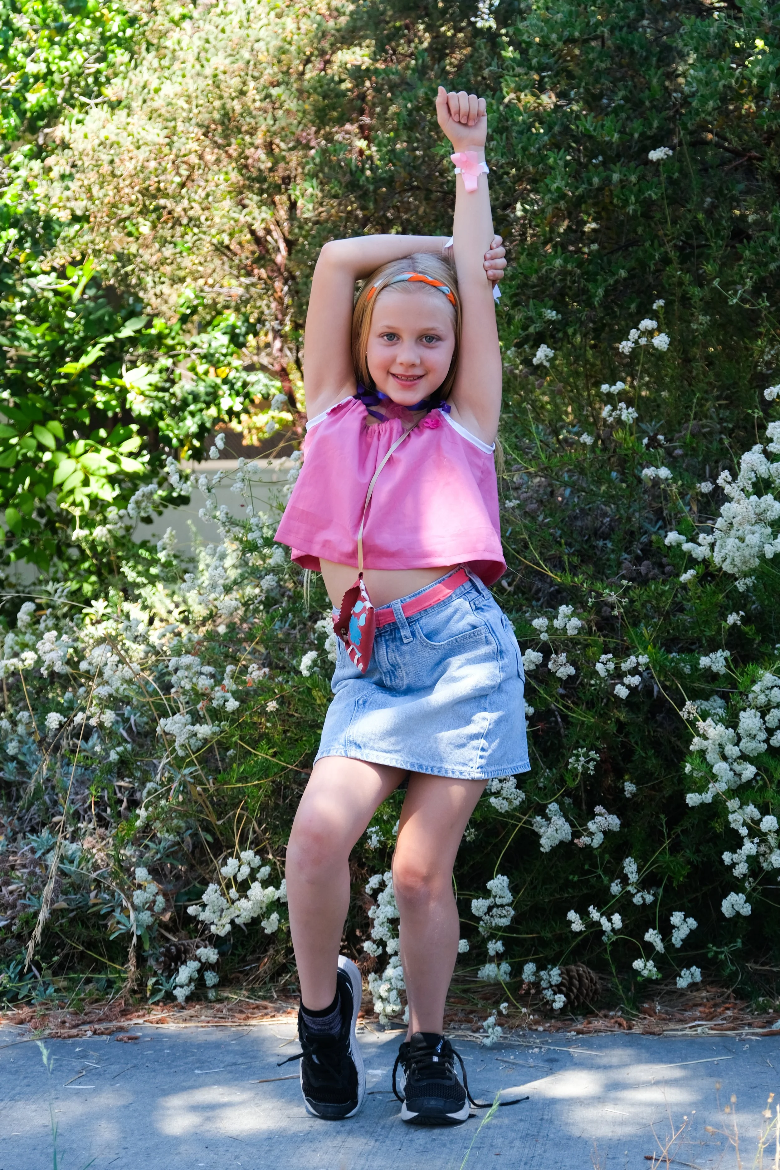 A young girl with blonde hair, wearing a pink top, denim skirt, and black sneakers, poses with arms raised outdoors in front of bushes with small white flowers.