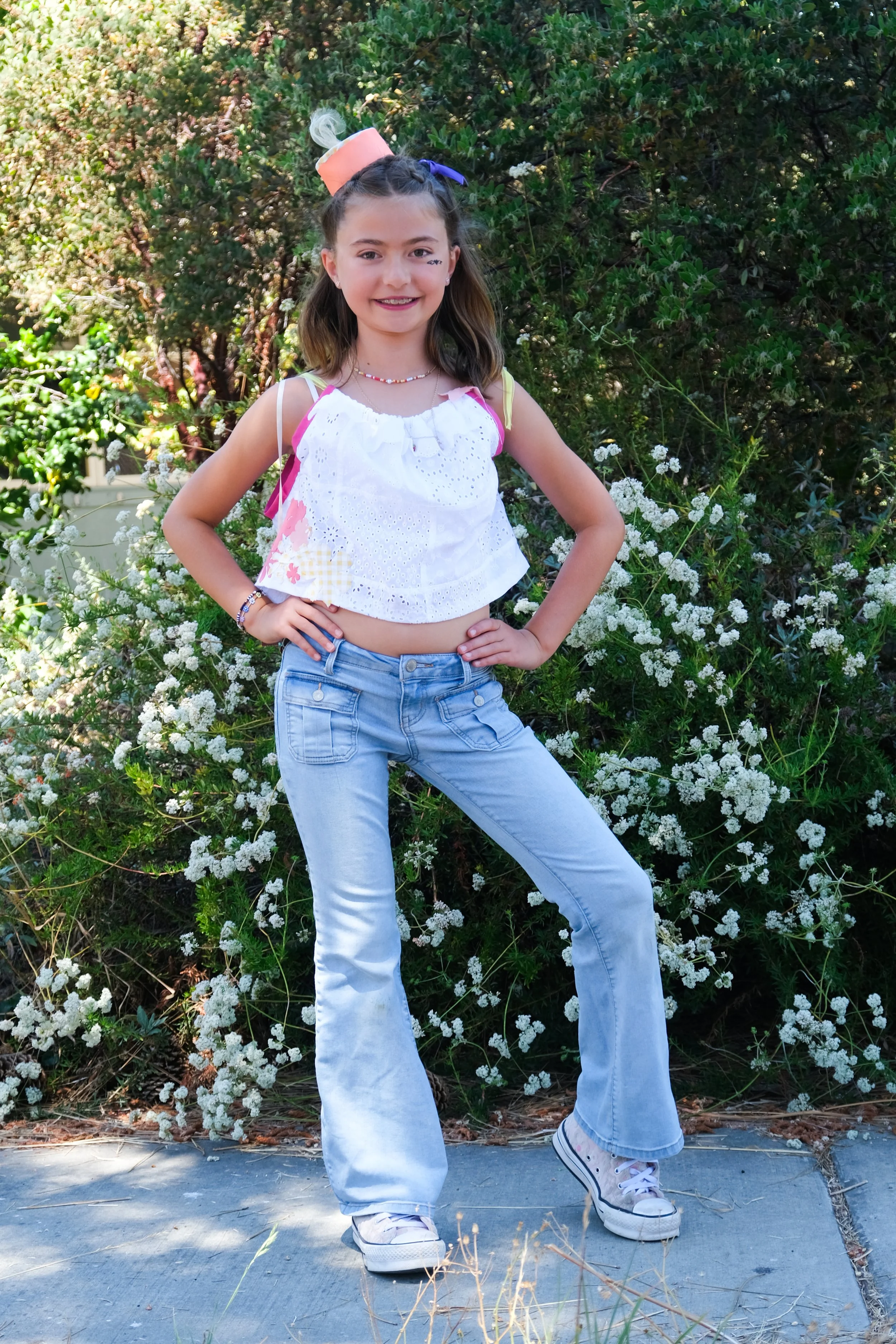 A young girl with long hair, wearing a white eyelet top, light blue jeans, and sneakers, standing outdoors in front of green foliage and white flowers, smiling at the camera with her hands on her hips.