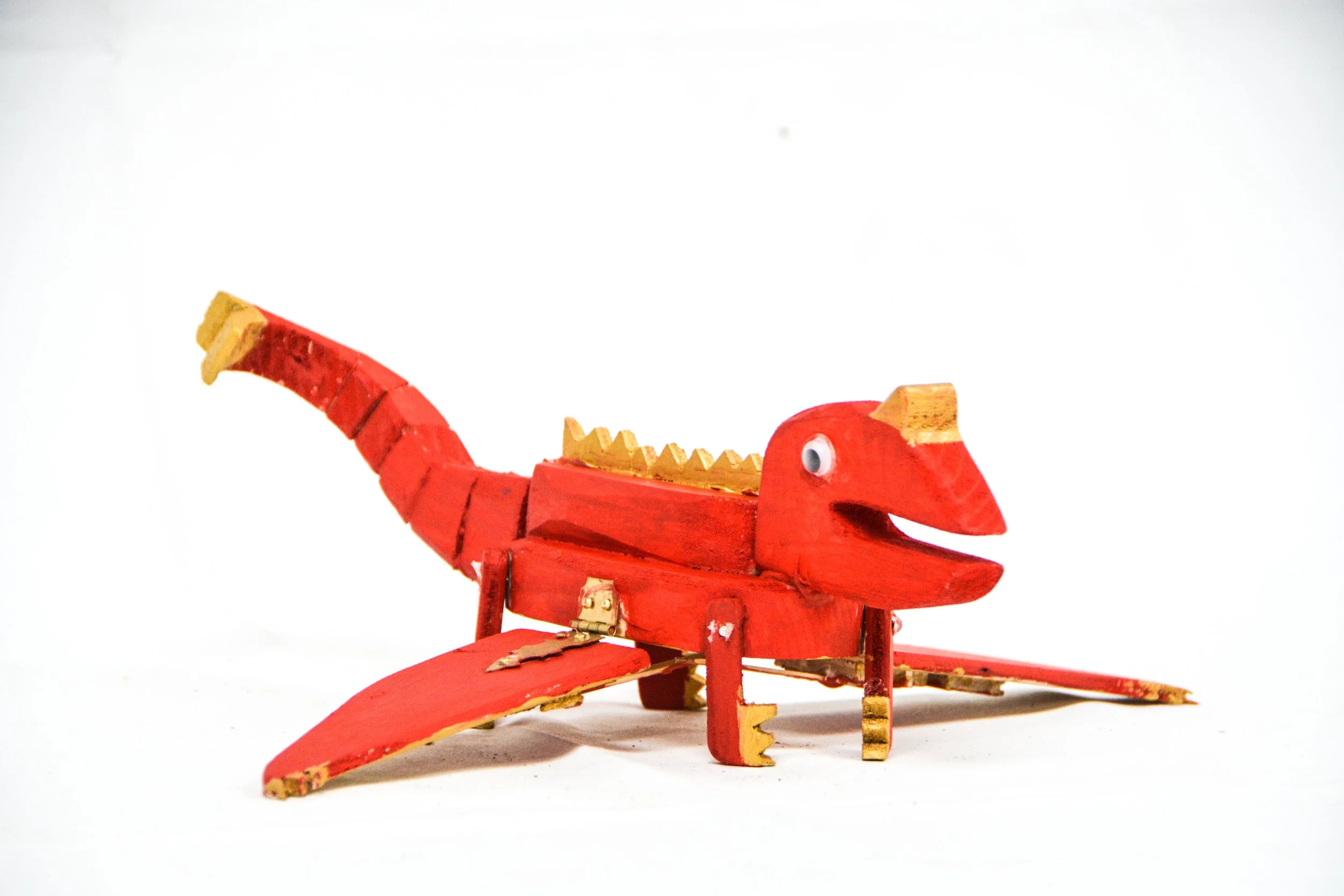 A red wooden toy dinosaur with painted details, googly eyes, and gold accents, placed on a plain white background.