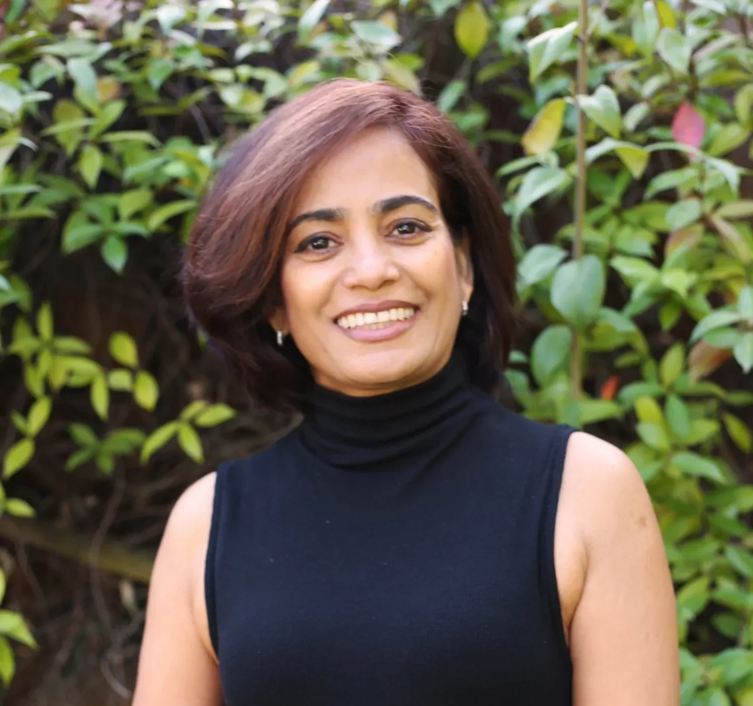 A woman with short brown hair smiling outdoors in front of green leafy bushes, wearing a black sleeveless turtleneck top.
