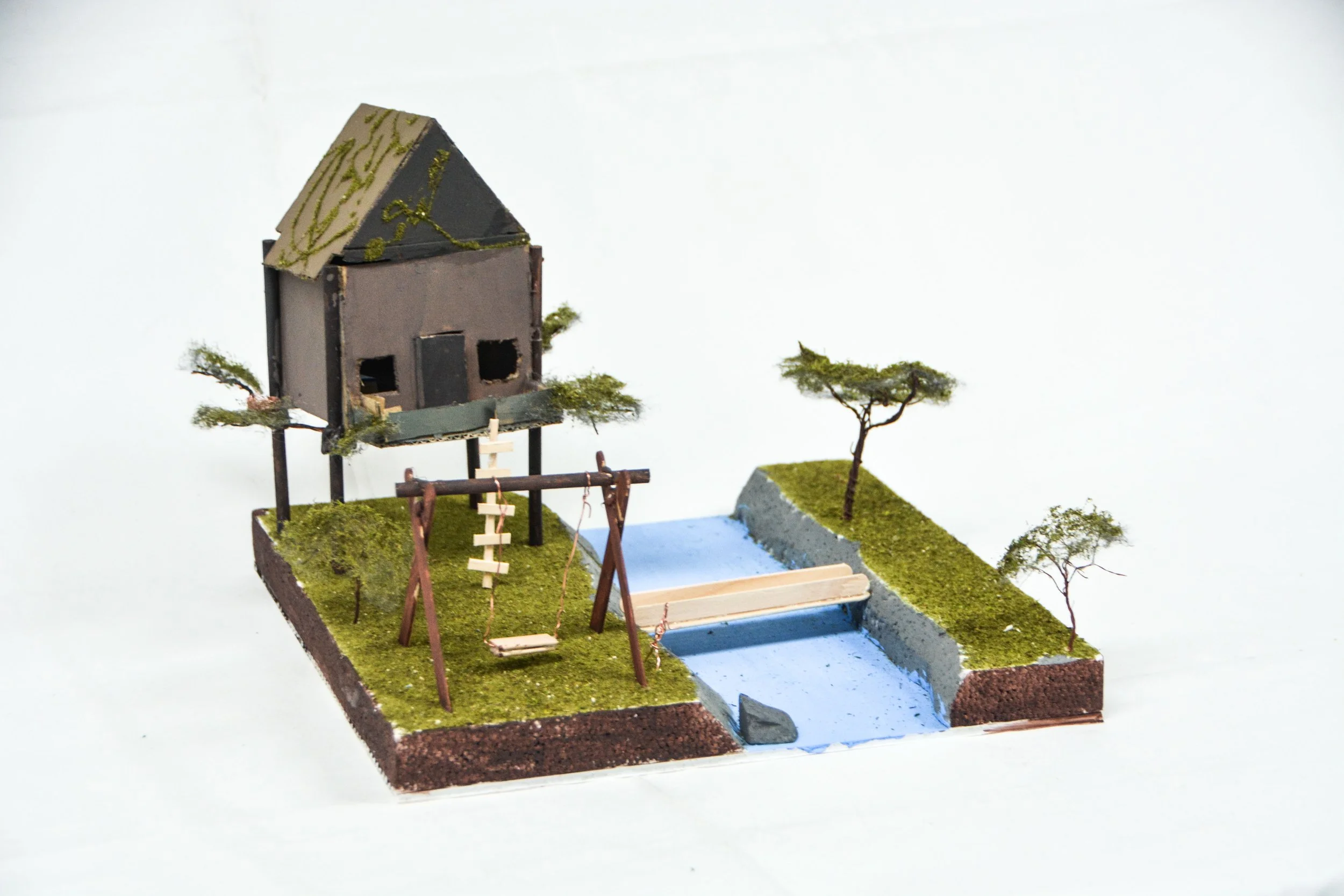 Miniature model of a house on stilts with a sloped roof, surrounded by green grass, small trees, a swing set, and a small pond.