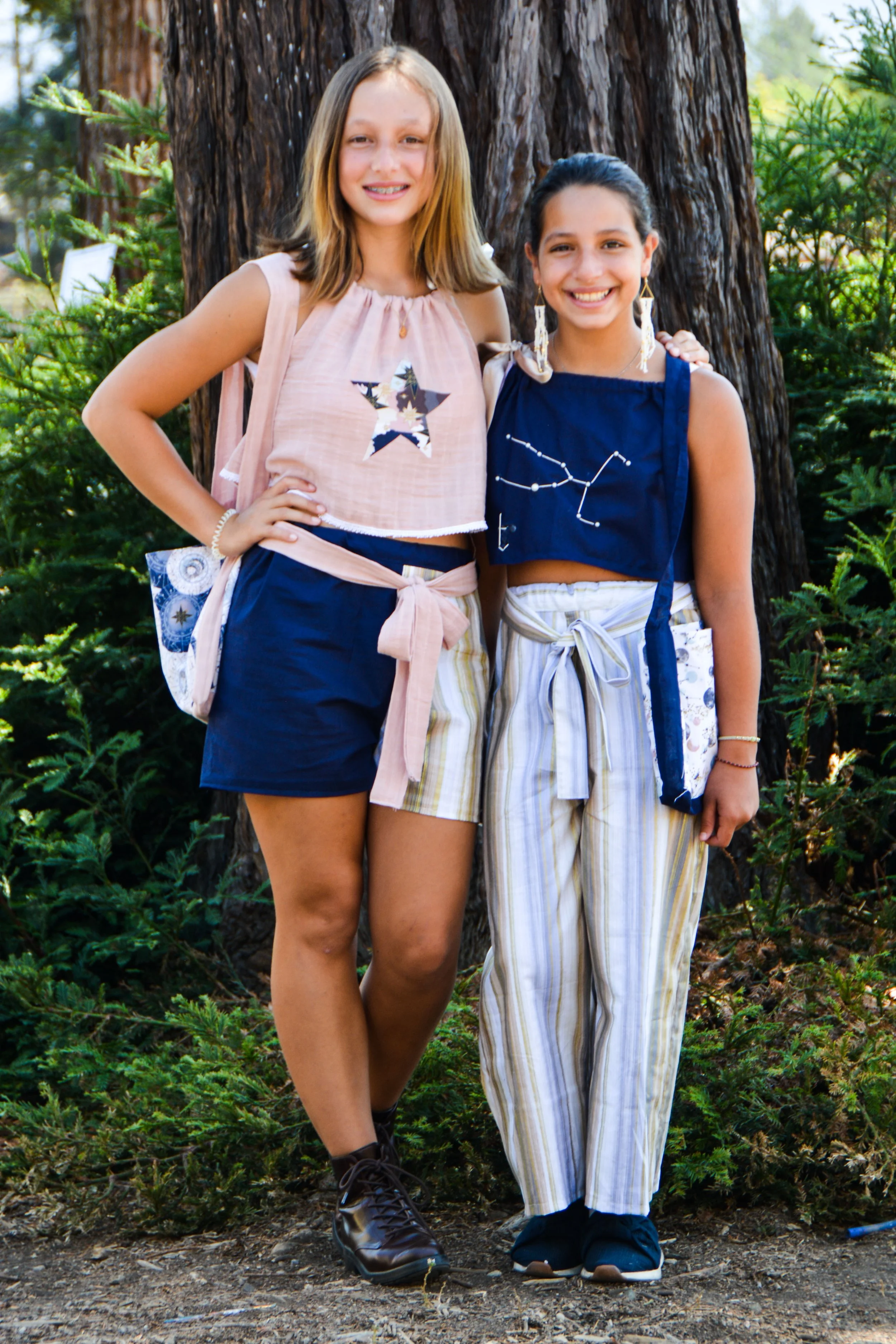 Two young girls standing outdoors in front of a tree, smiling at the camera. One girl has shoulder-length blonde hair, wearing a light pink sleeveless top with a star design, navy shorts, and black shoes. The other girl has long dark hair, wearing a 