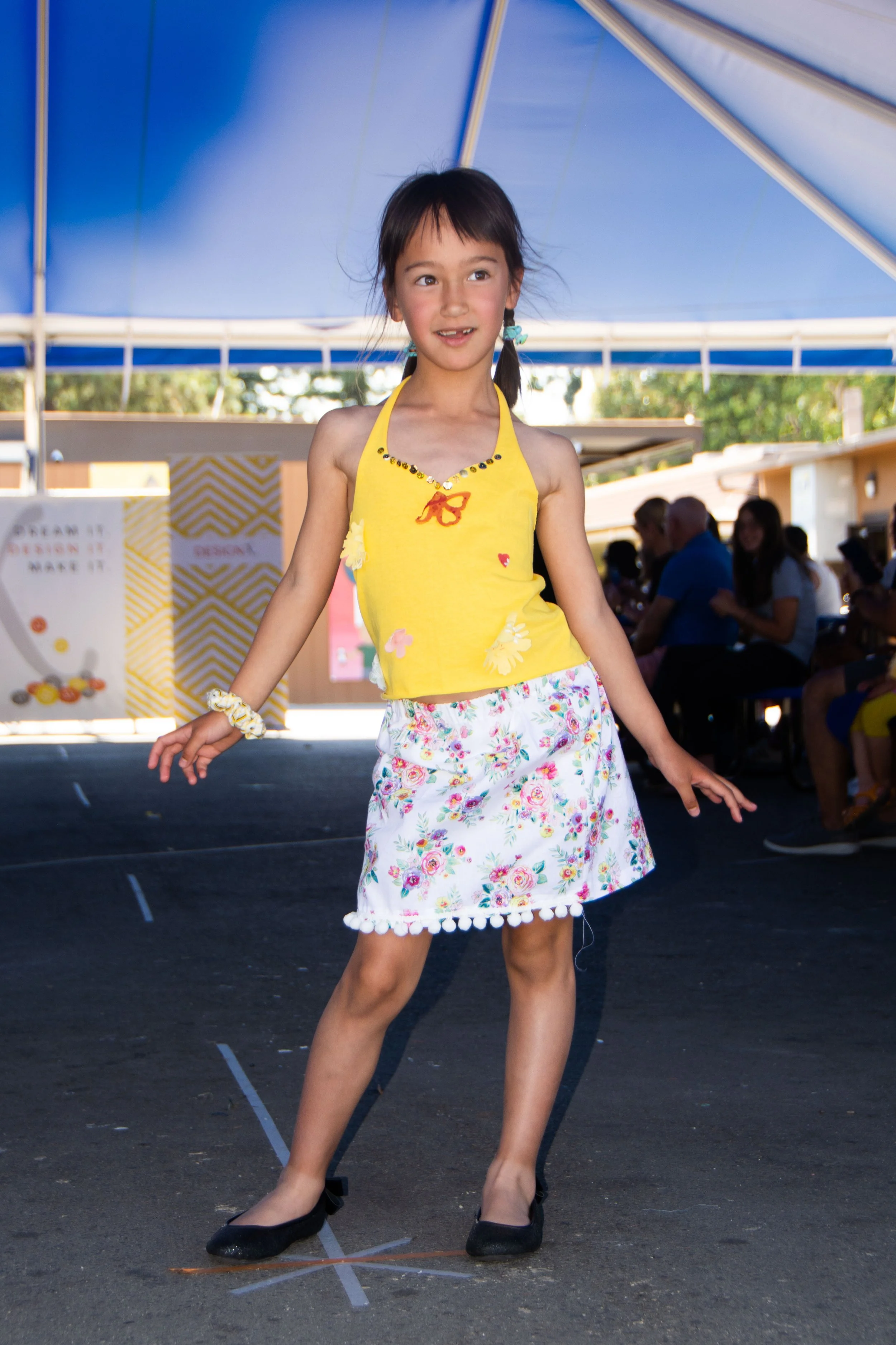 Young girl with dark hair in pigtails, wearing a yellow sleeveless top with floral decorations, a white floral skirt, and black shoes, dancing or performing on an outdoor stage under a blue canopy with an audience in the background.