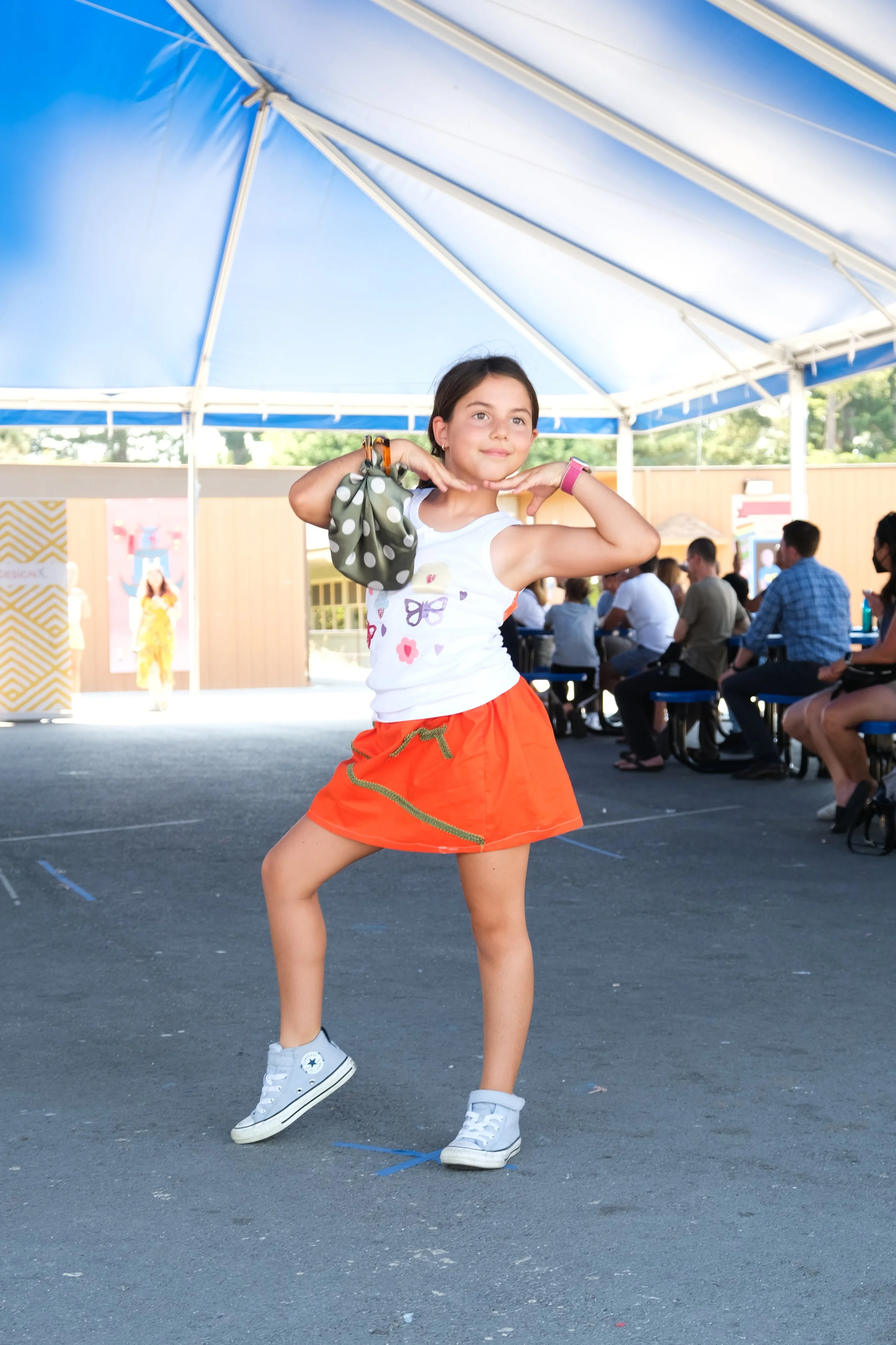 A young girl dancing on an outdoor stage with an audience seated in the background under a blue and white canopy.