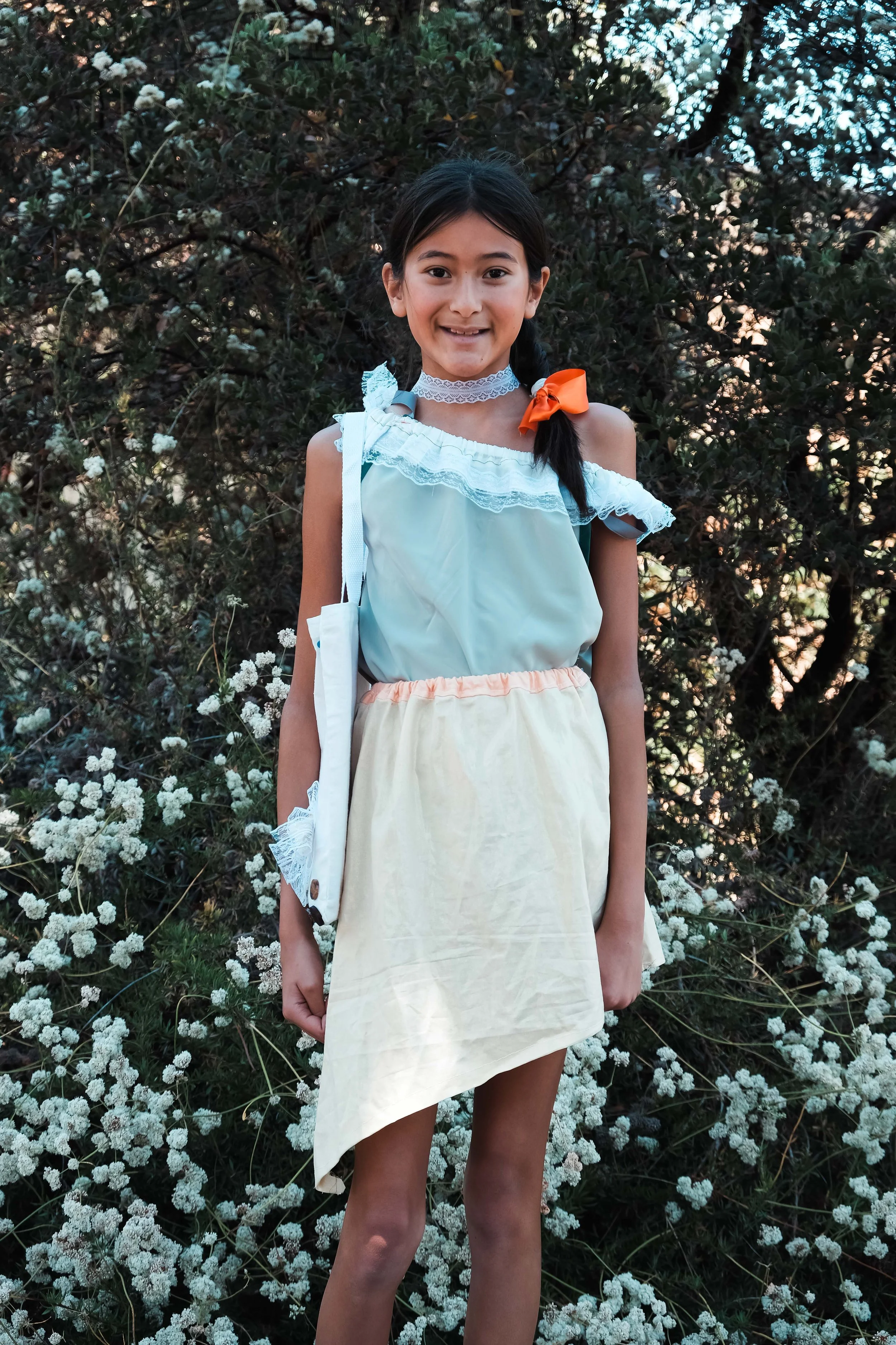 A young girl with long dark hair in pigtails, dressed in a colorful outfit, stands outdoors in front of flowering bushes. She is smiling and facing the camera.