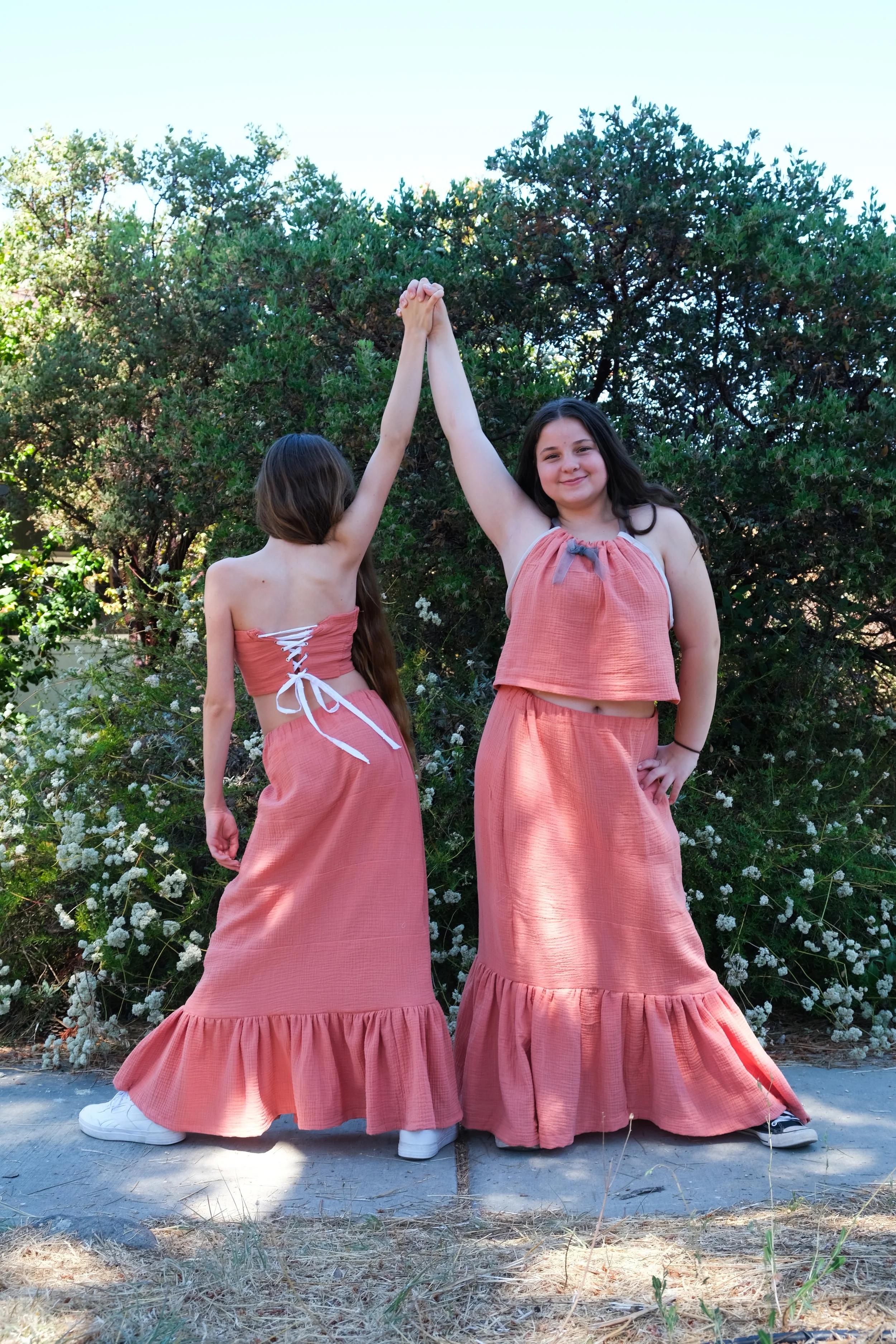 Two young girls in matching coral-colored ruffle skirts and tops holding hands up and smiling outdoors with greenery in the background.