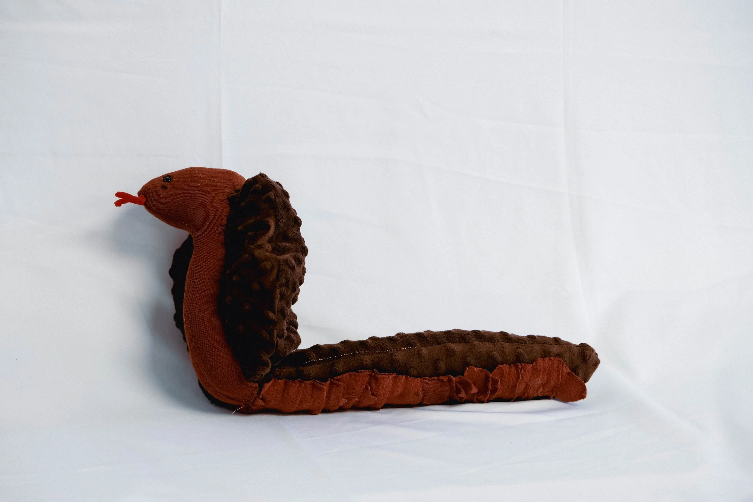 Soft plush toy in the shape of a serpent, with a brown and orange body, black textured dorsal fin, and a small red tongue, placed on a plain white surface.
