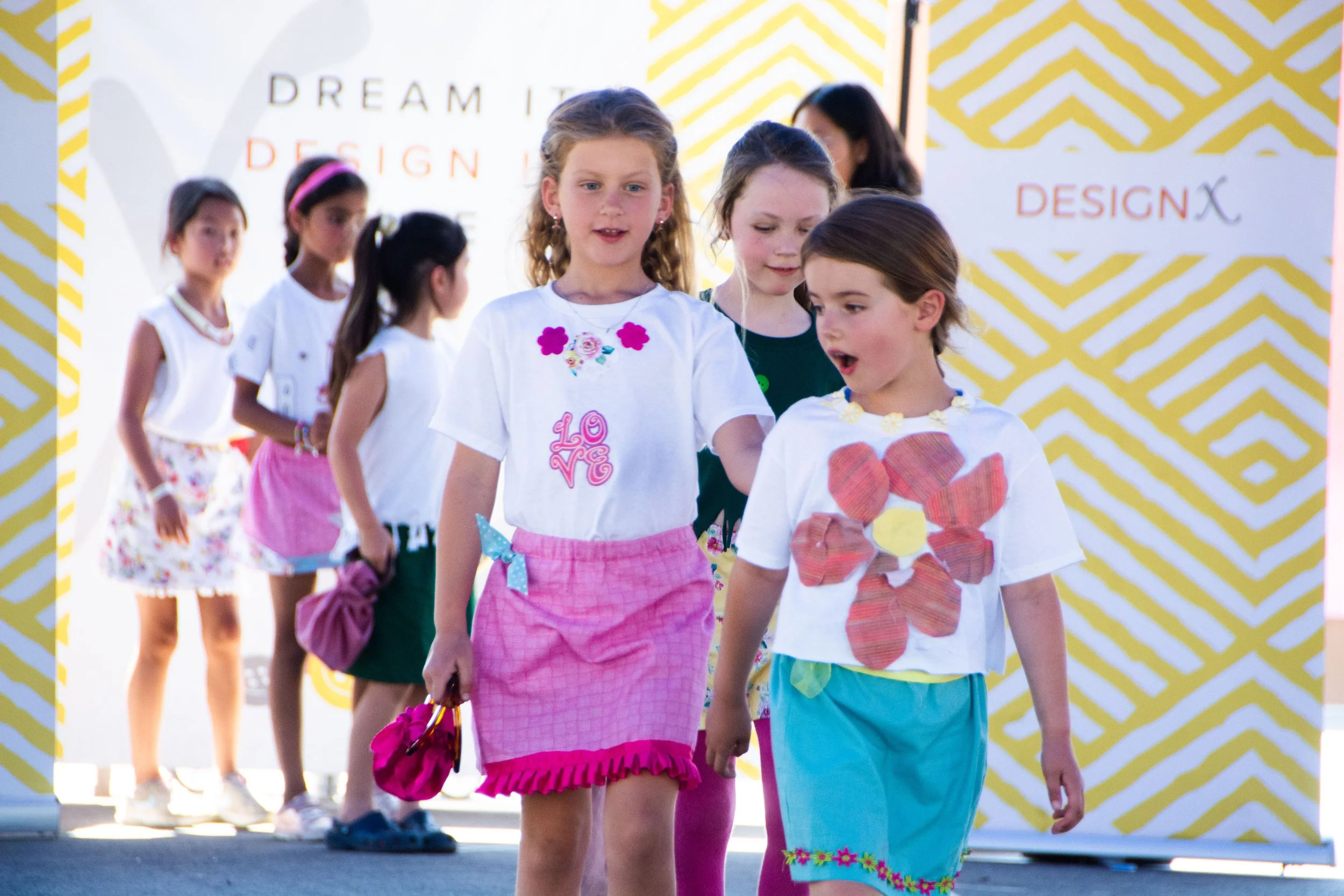 Group of young girls walking in a fashion show, wearing colorful outfits with floral and love designs, on a runway with a yellow and white zigzag patterned background.
