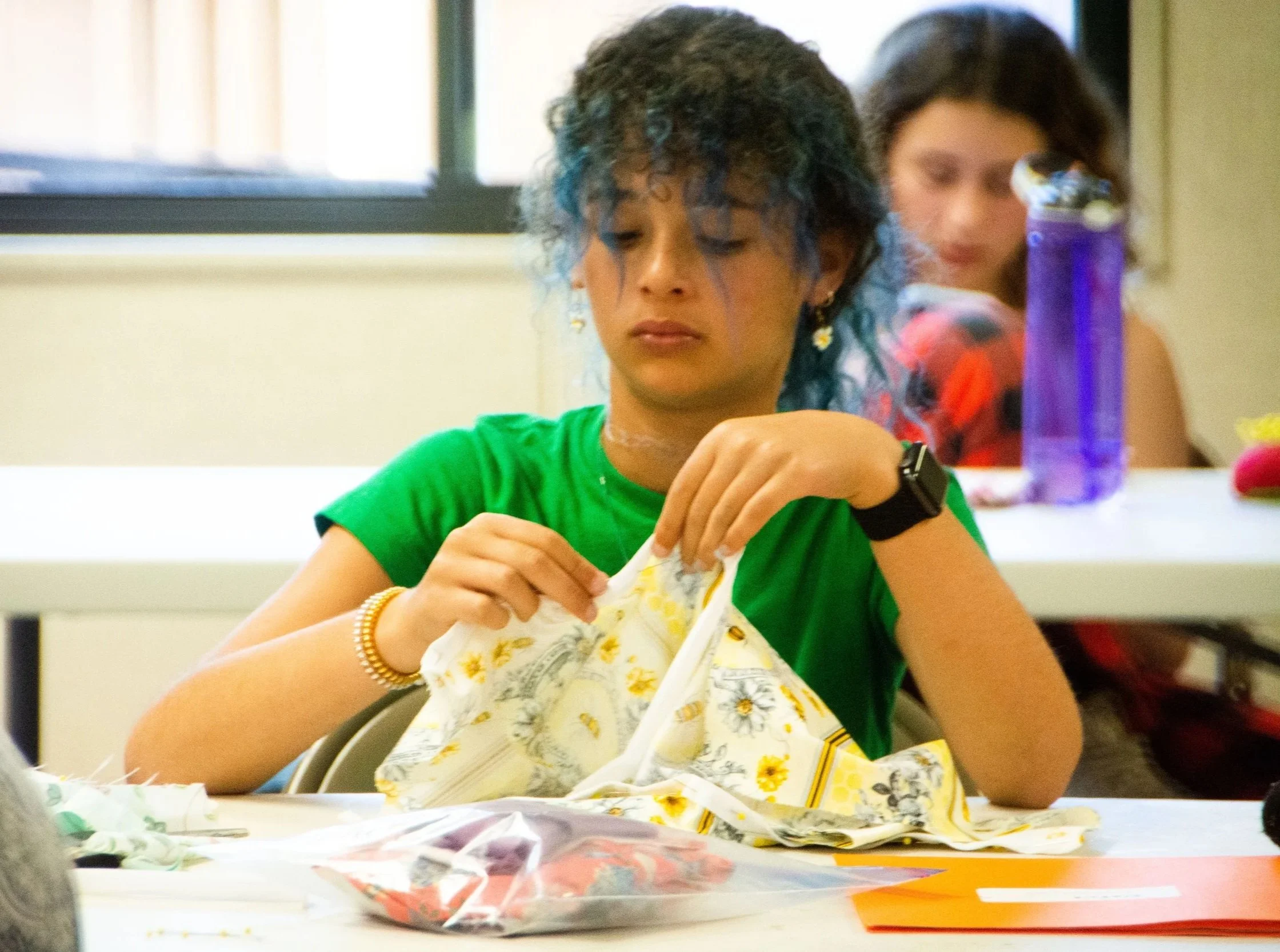 A young DesignX fashion student is sewing a piece of fabric with a yellow and white floral pattern. Another girl with dark hair and a red shirt is in the background, with a purple water bottle on the table in front of her.