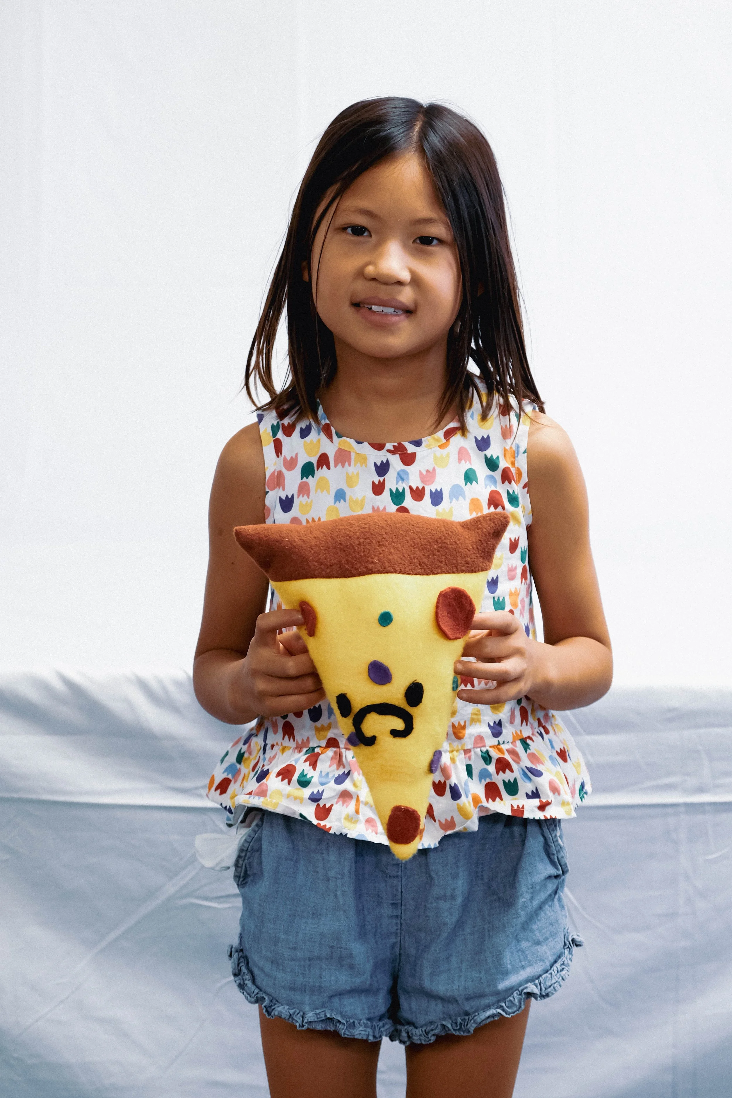 A young girl with shoulder-length dark hair in a colorful, patterned sleeveless top and denim shorts holding a plush pizza slice with an animated face, standing against a plain background.