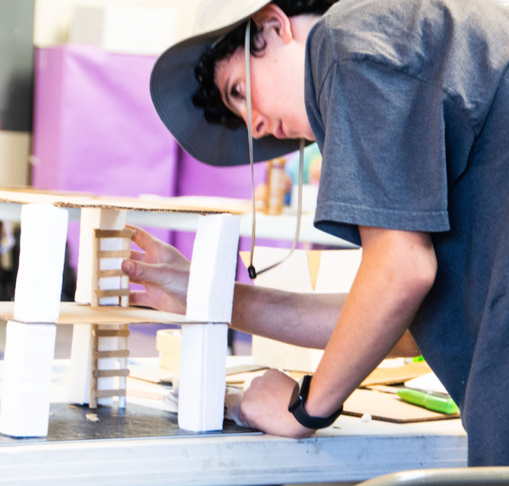 A young person wearing a wide-brimmed hat working on a small architectural model made of wooden sticks and foam board.