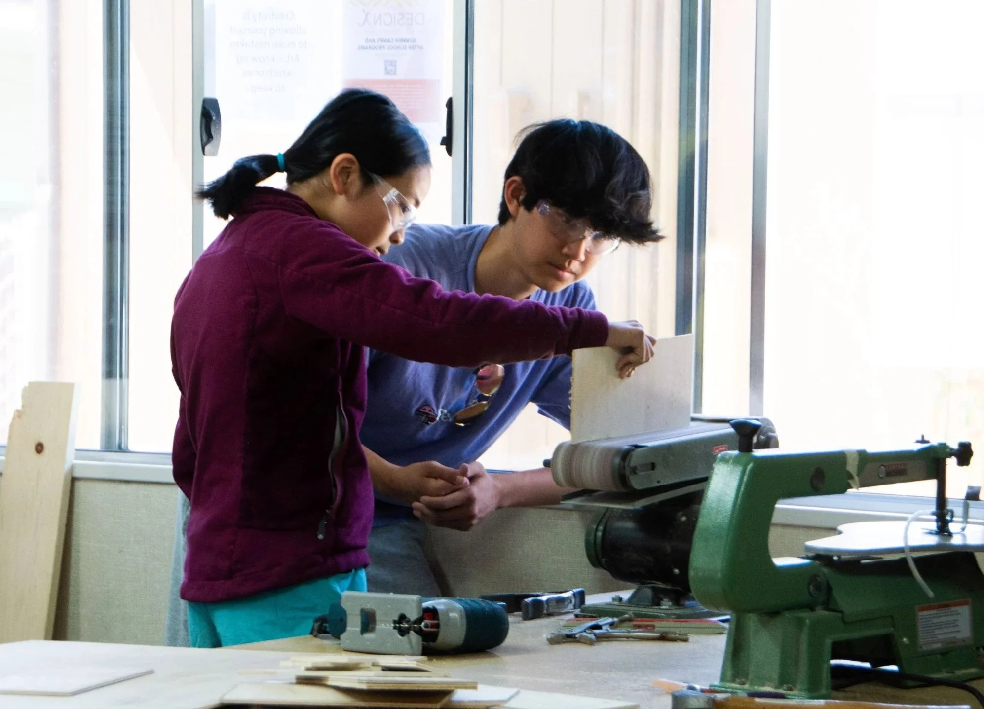 Two people working together in a woodworking shop, cutting a piece of wood using a large green woodworking machine near a window.
