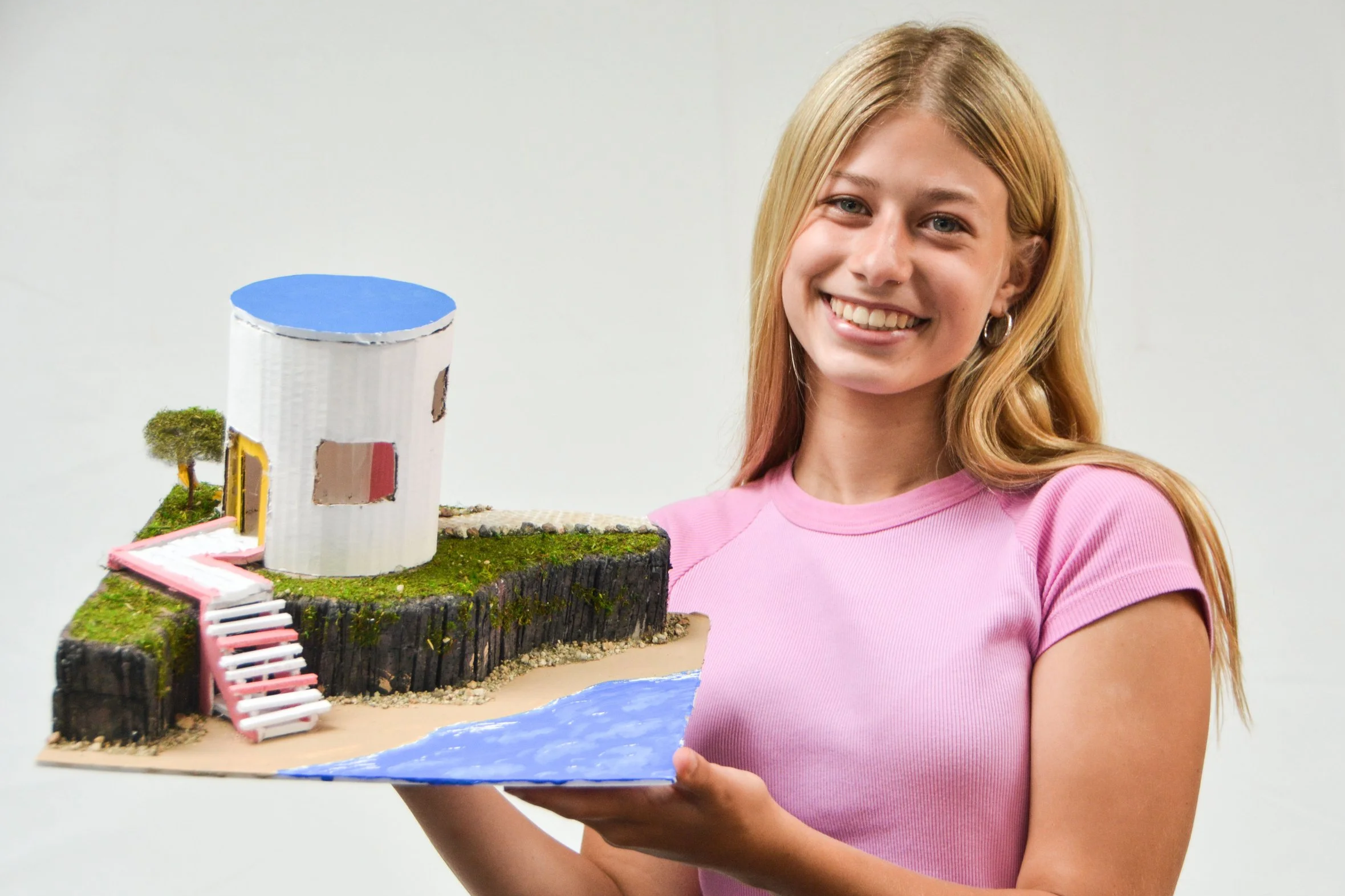 A woman with blonde hair wearing a pink shirt holding a miniature model of a building with a cylindrical tower next to water.
