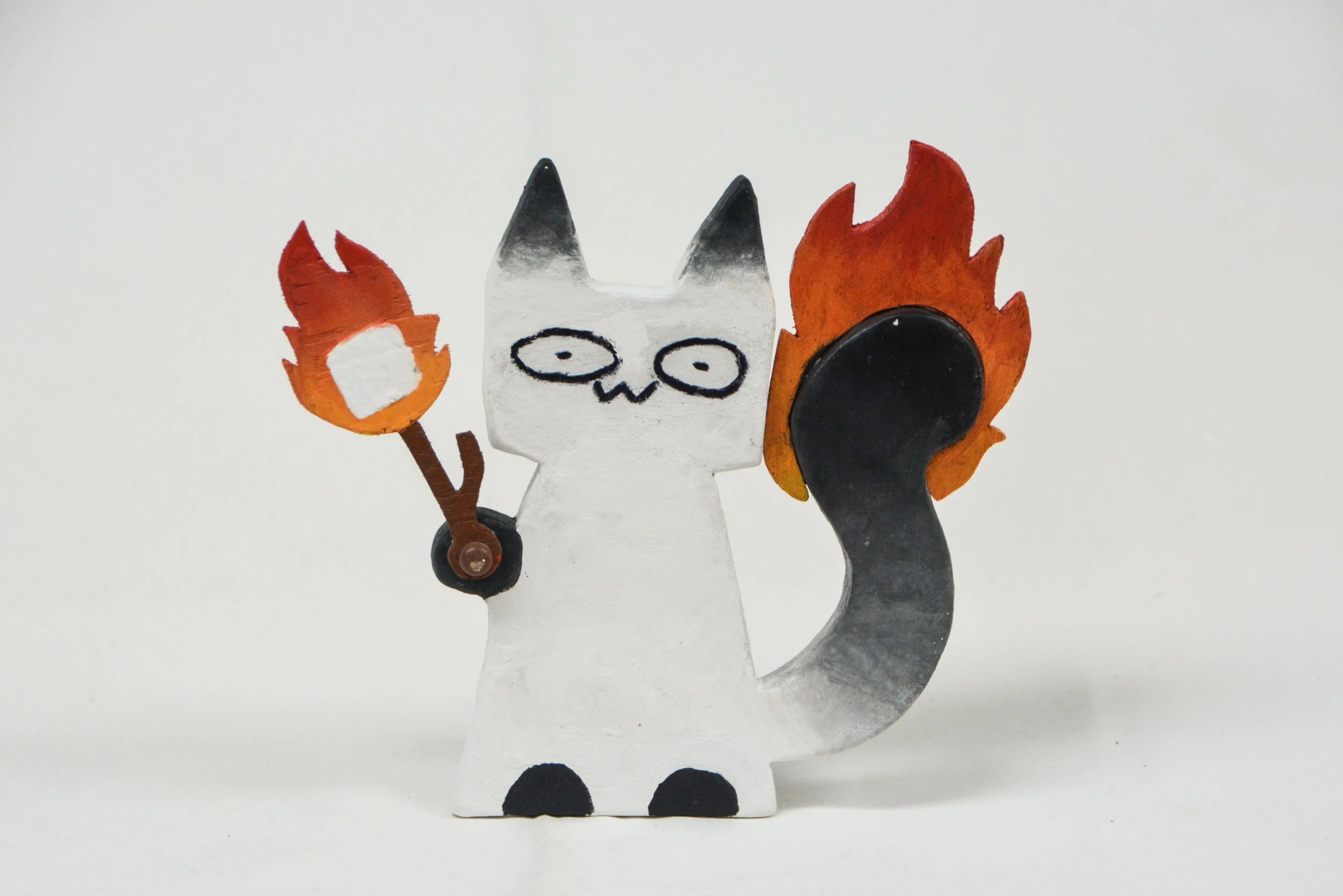 Handmade figurine of a white cat with black ears, holding a flaming torch in one paw, with a tail on fire. The cat has simple facial features, including round eyes and a small mouth.