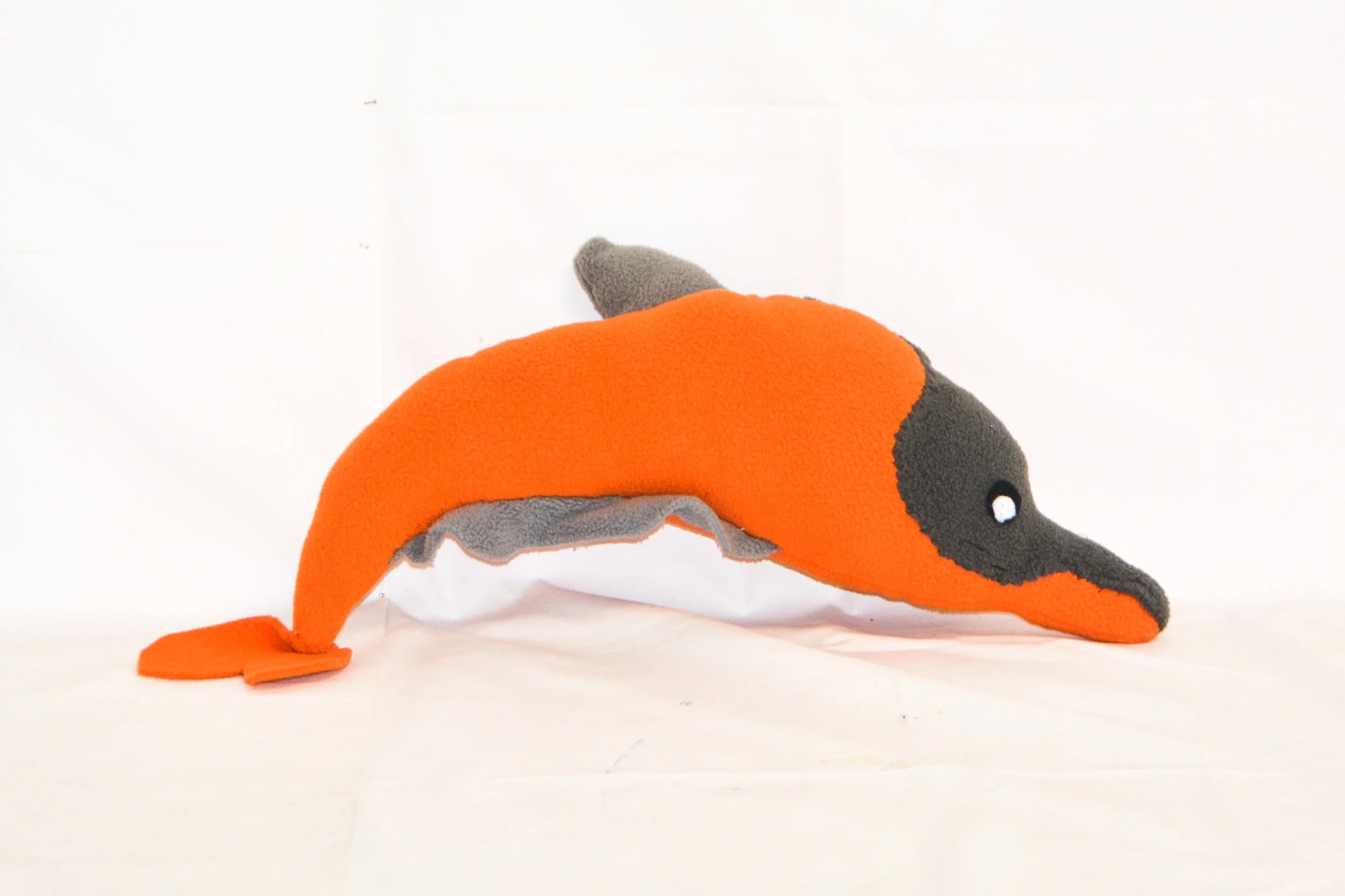 A stuffed toy resembling an orange and gray narwhal with a long, pointy nose and a small tail, set against a plain white background.