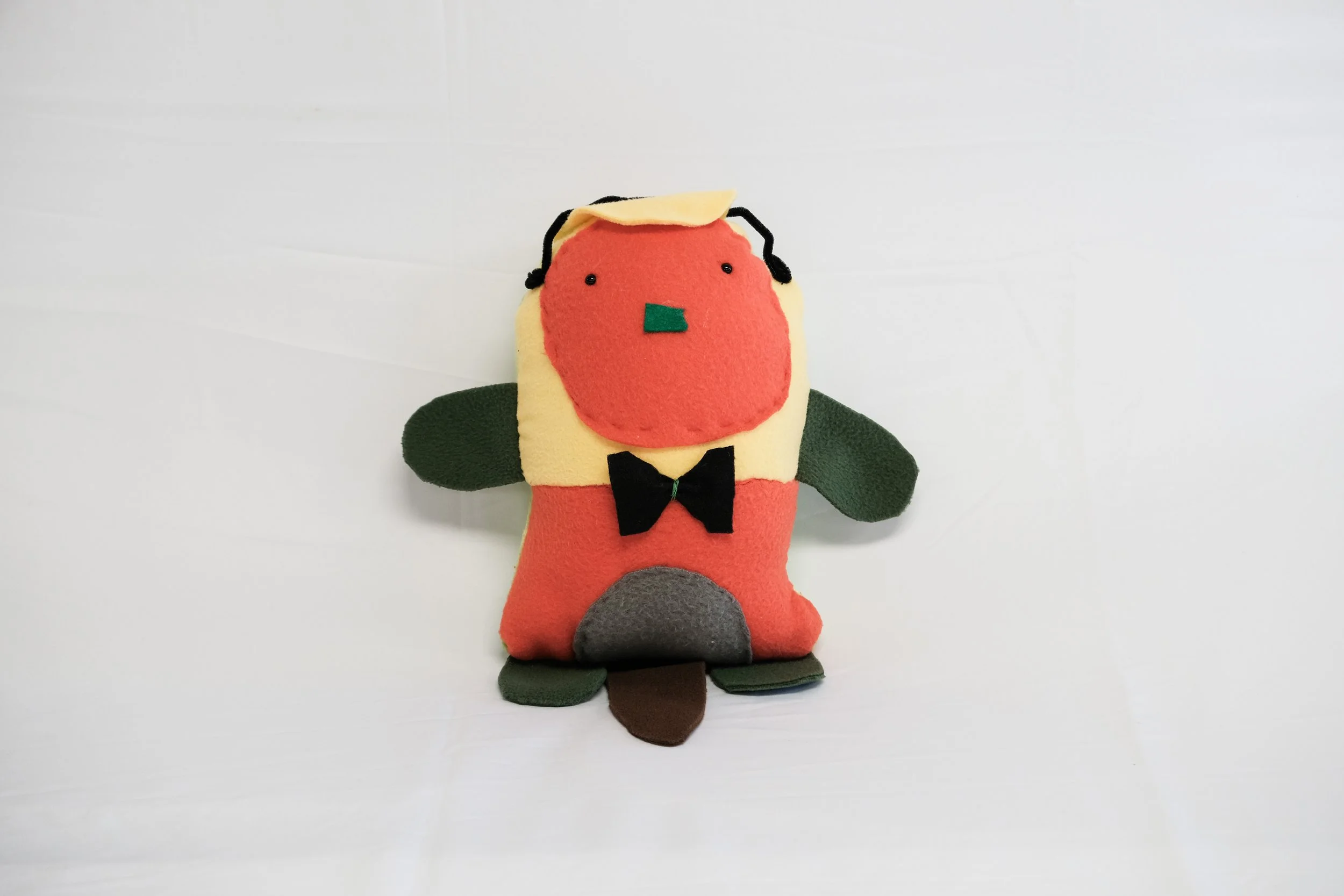 A plush toy designed to resemble a colorful animal character with a red face, black eyes, and a blue mouth. It has green ears, a black bow tie, and a grey patch on its belly, standing on a white surface.