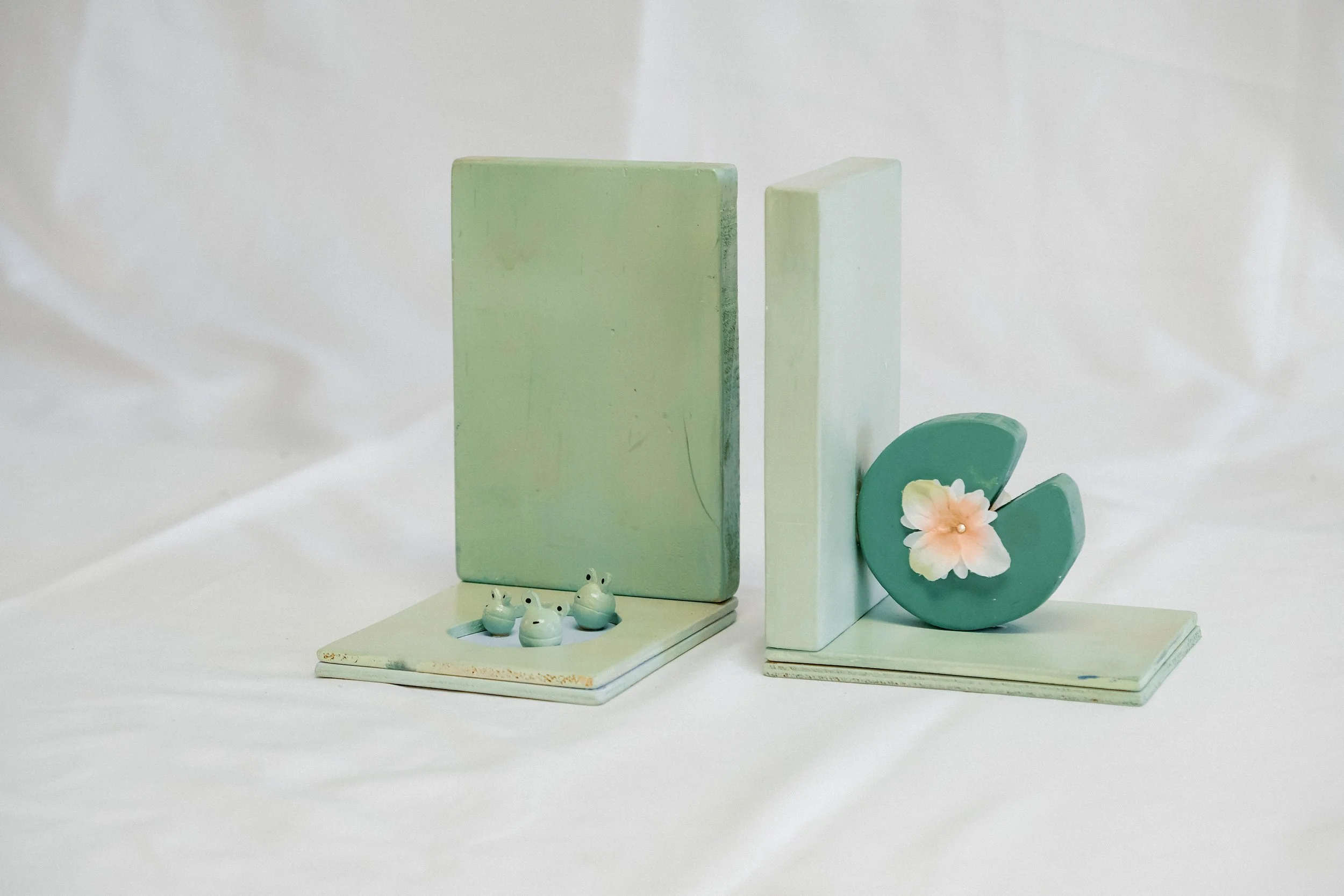 Decorative pastel-colored bookends with frog figurines and a flower on a white fabric background.