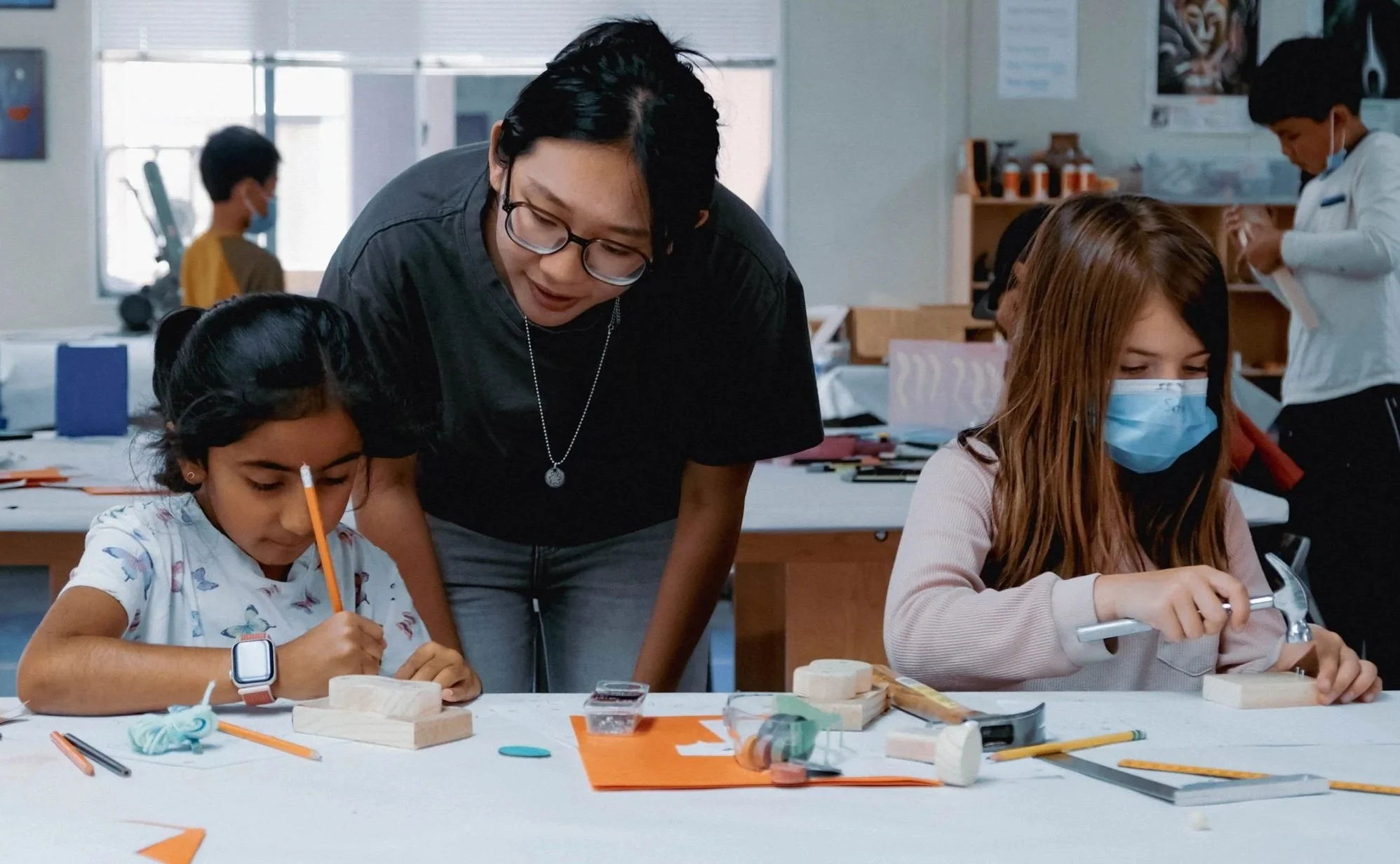 Two young girls wearing face masks working on woodworking projects at a table, with a young woman assisting them in a classroom setting.