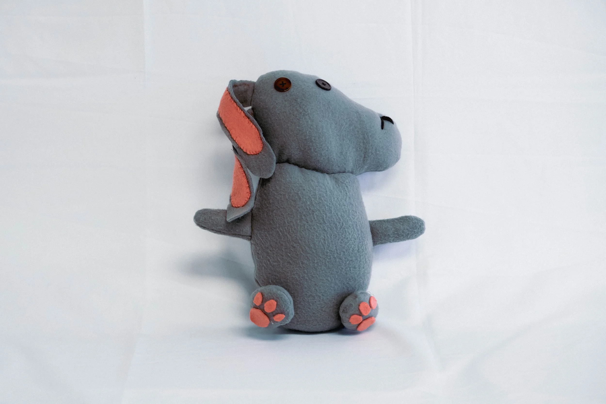 Grey plush stuffed animal resembling a dog with pink accents on ears and paws, sitting against a white background.