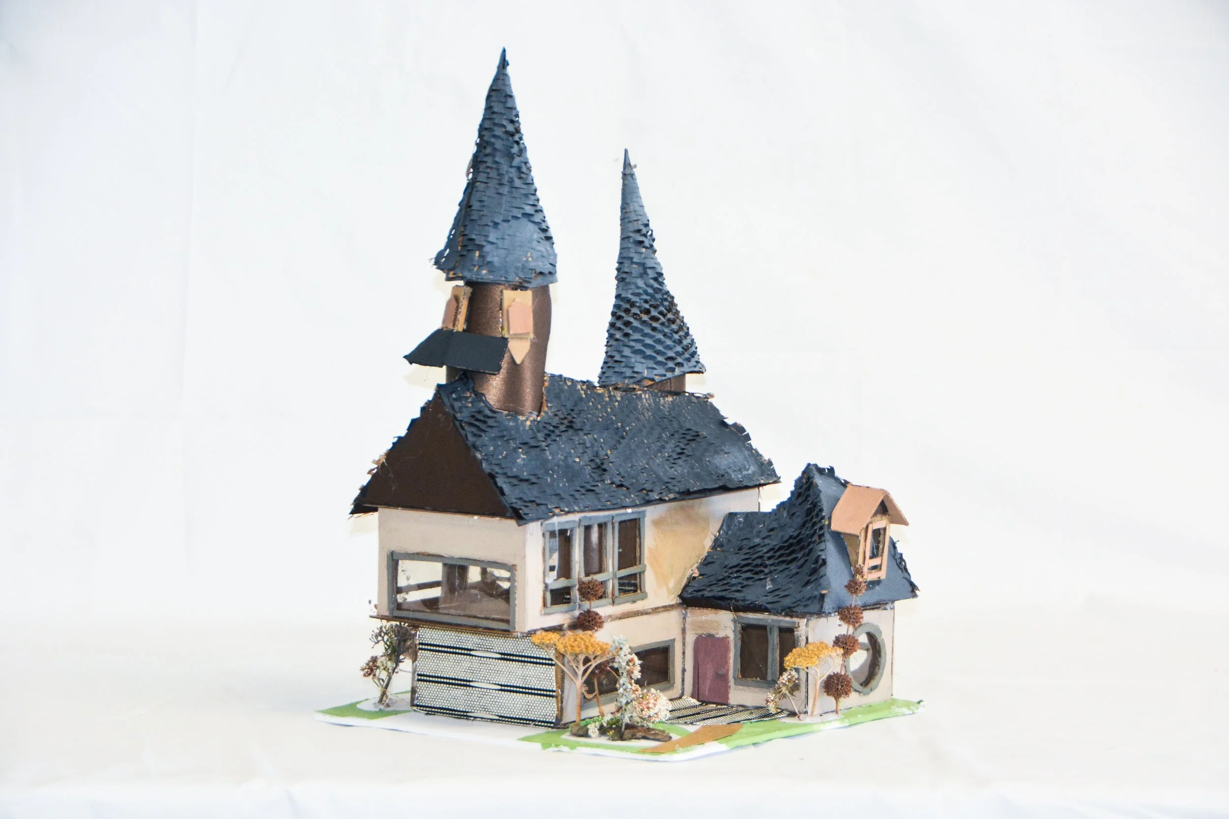 A miniature model of a whimsical house with two tall, pointed, blue-tiled towers on a white background. The house features a large front window and small trees with autumn-colored leaves around it.