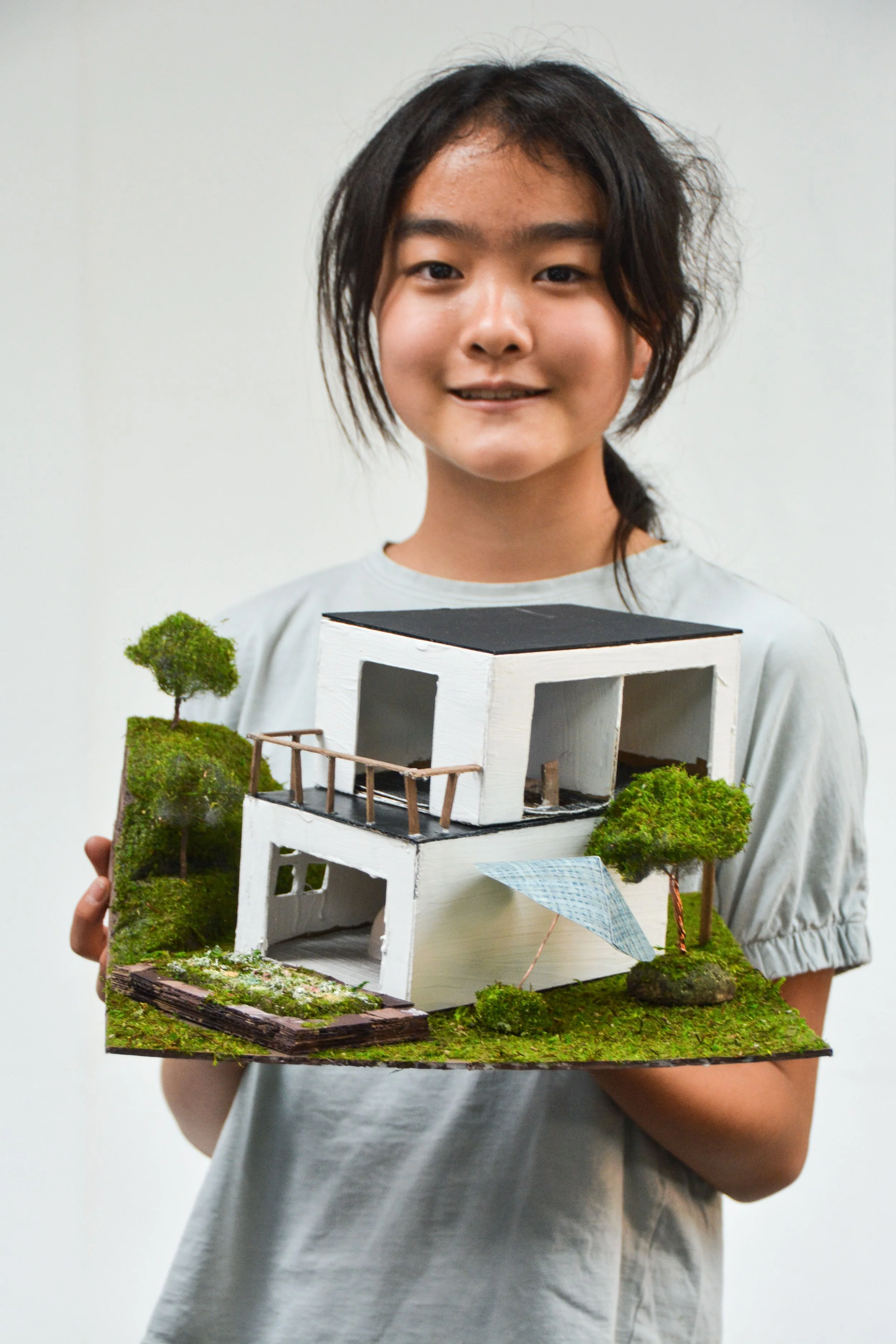 Young girl holding a detailed architectural model of a modern house with trees, moss, and miniature landscaping.