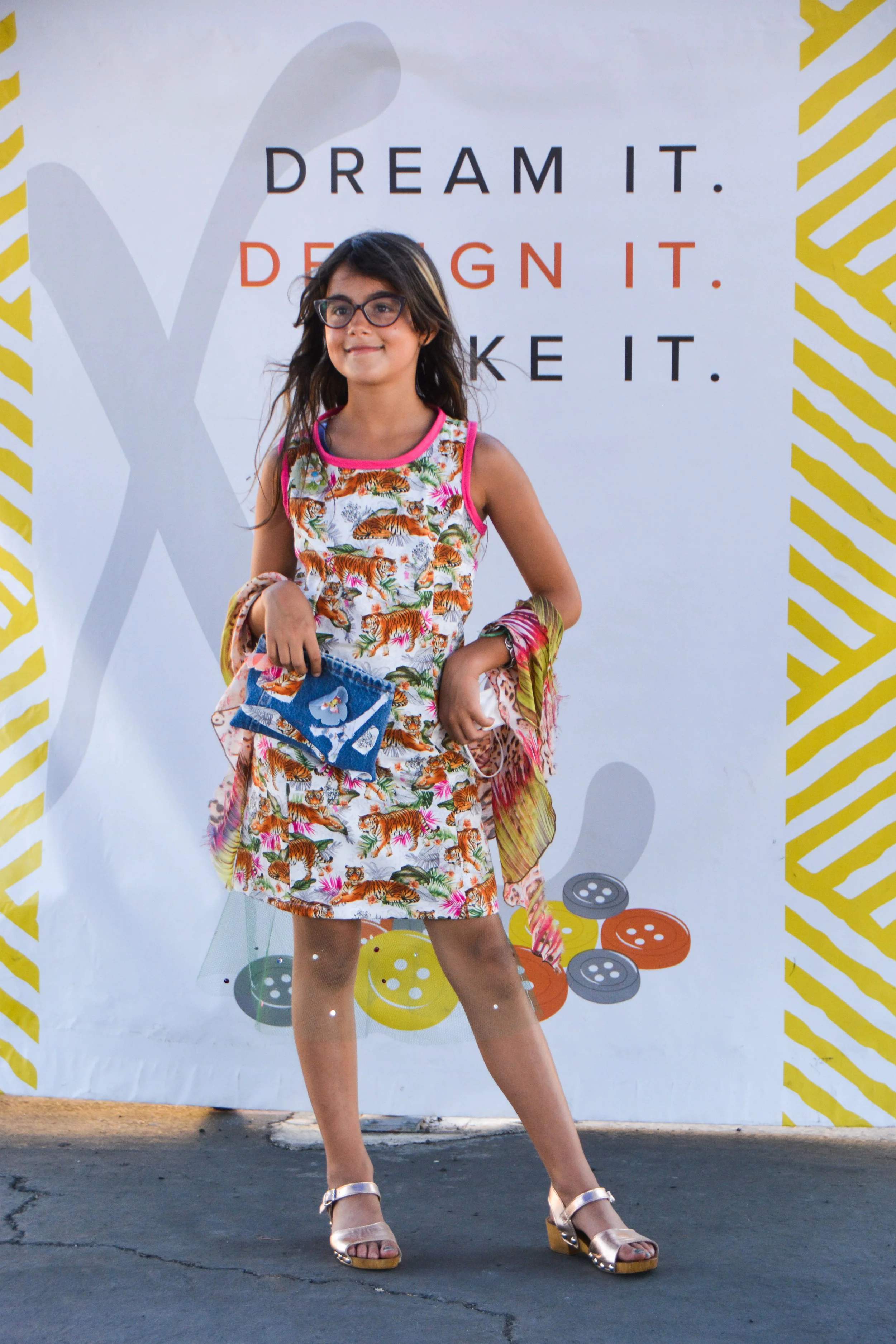 A girl wearing glasses and a colorful dress with a tiger print, standing in front of a DesignX backdrop with the words 'Dream It. Design It. Make It.' and a pattern of yellow stripes and buttons.