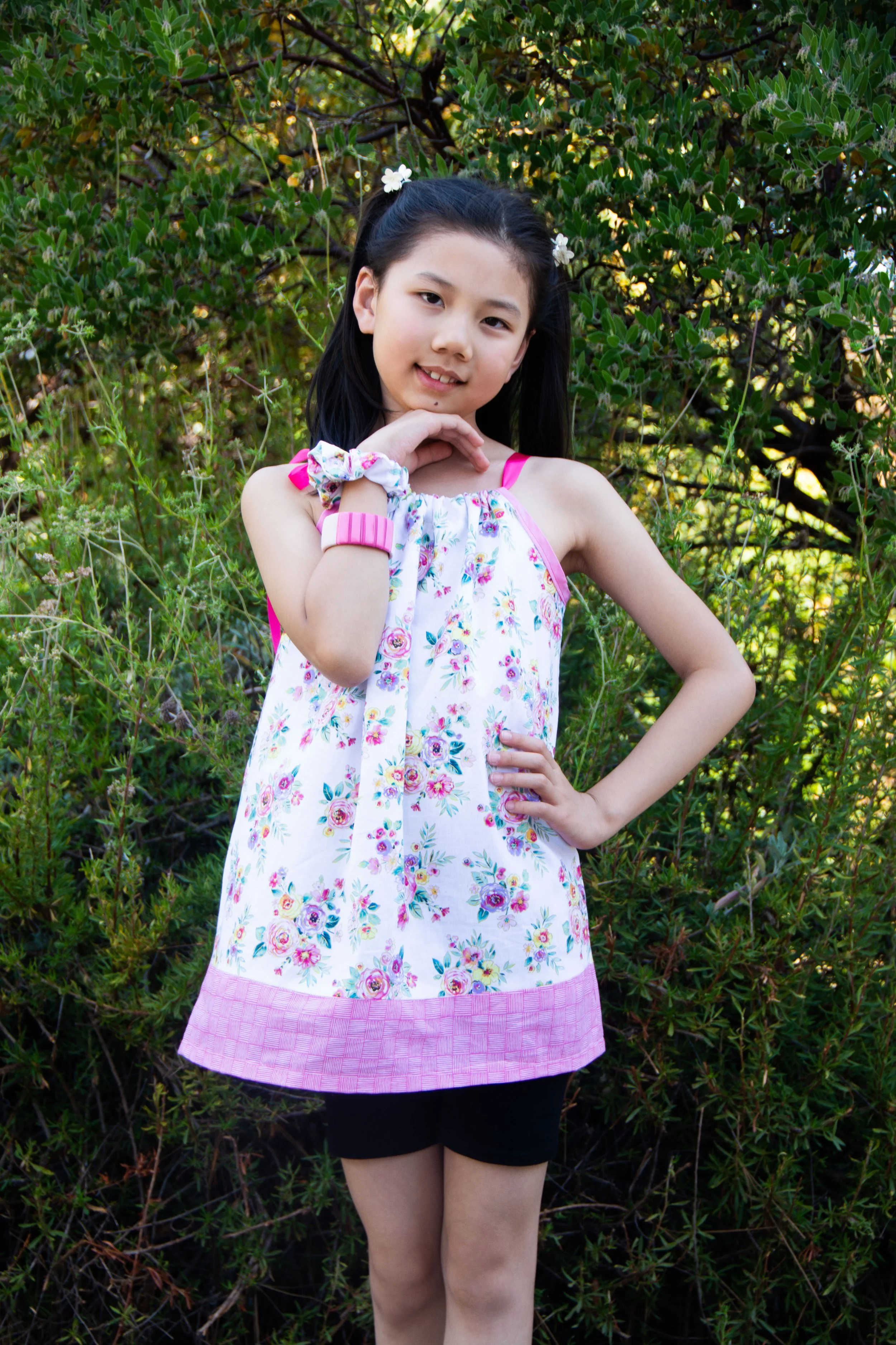 Young girl wearing a floral sleeveless dress with pink straps, black shorts, and a pink wristband, standing outdoors near green bushes, with her right hand on her hip and her left hand supporting her chin, looking at the camera.
