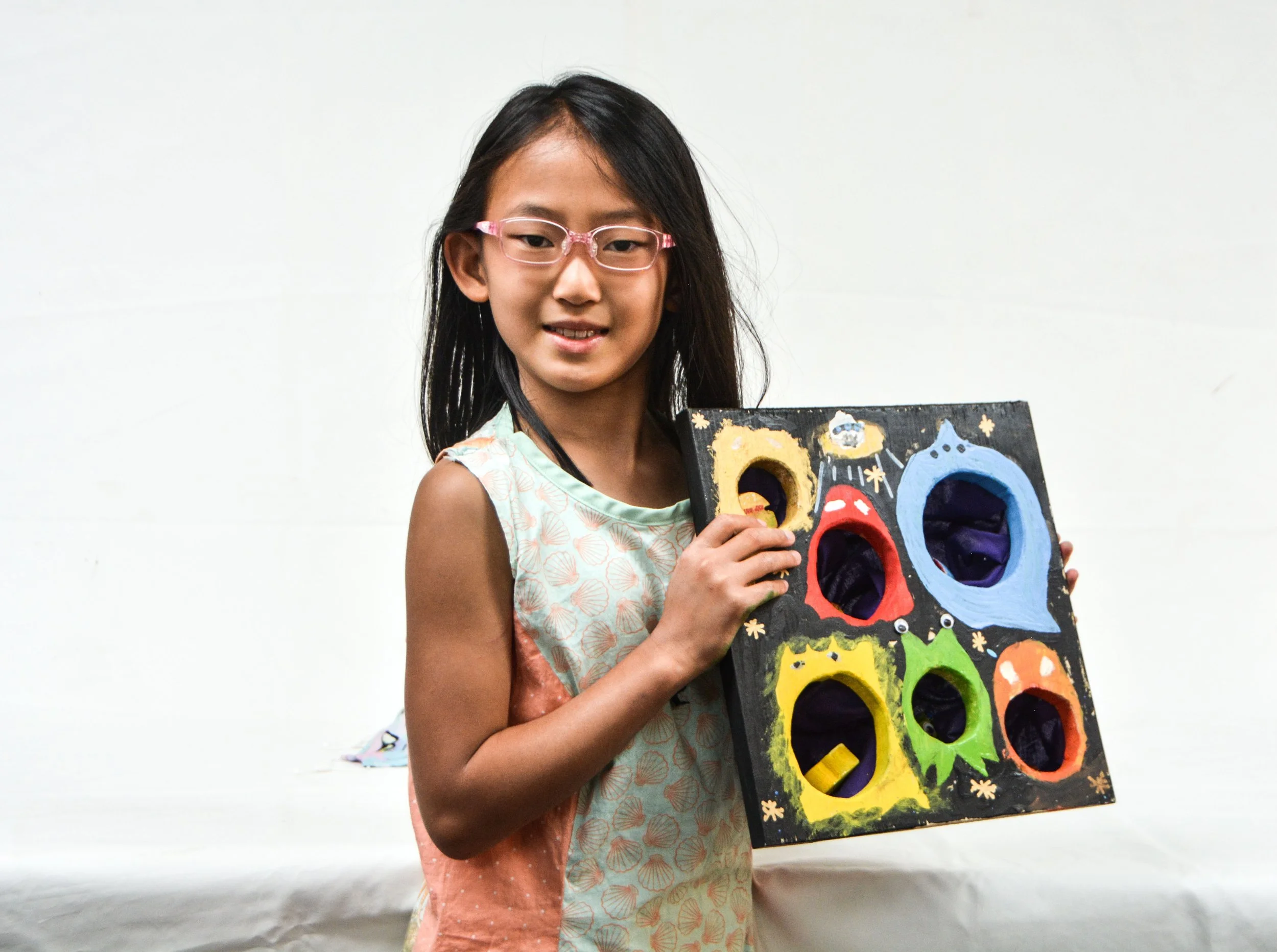 A young girl with glasses holding a colorful handmade board game with holes and monster faces painted on it.