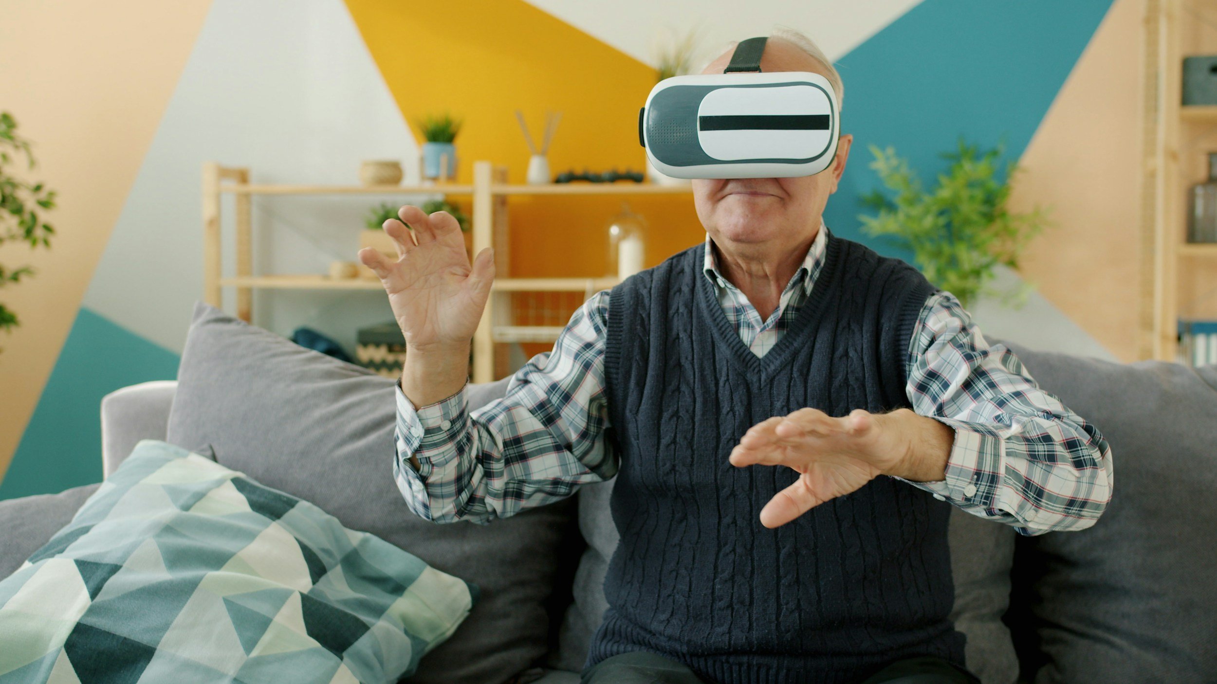 An elderly man wearing a virtual reality headset, sitting on a couch in a colorful living room, making gestures with his hands.