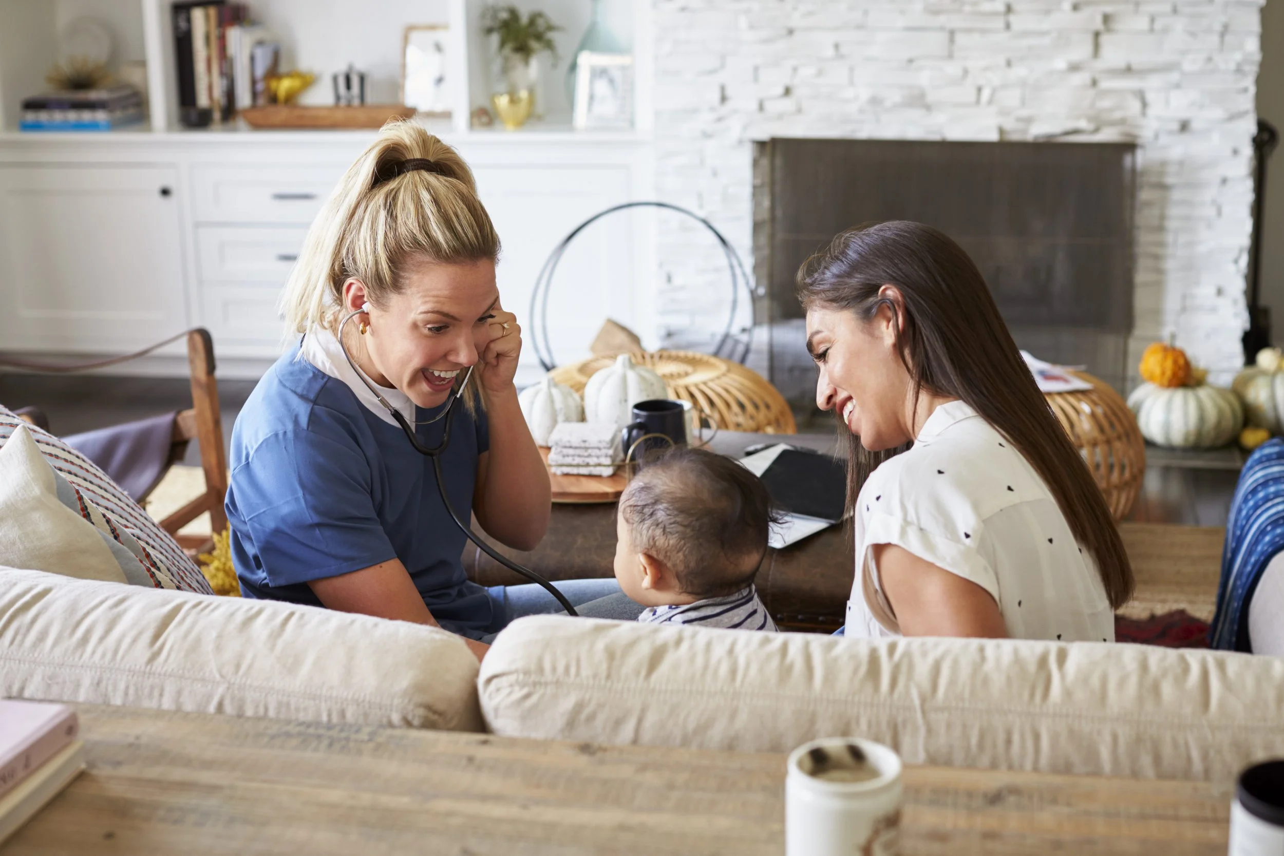 A woman in medical scrubs, holding a stethoscope to her ear, smiling and playfully listening to a baby sitting on a sofa. Another woman sitting beside the baby is smiling at the scene. The setting is a cozy living room with decorative pumpkins on a table in the background.