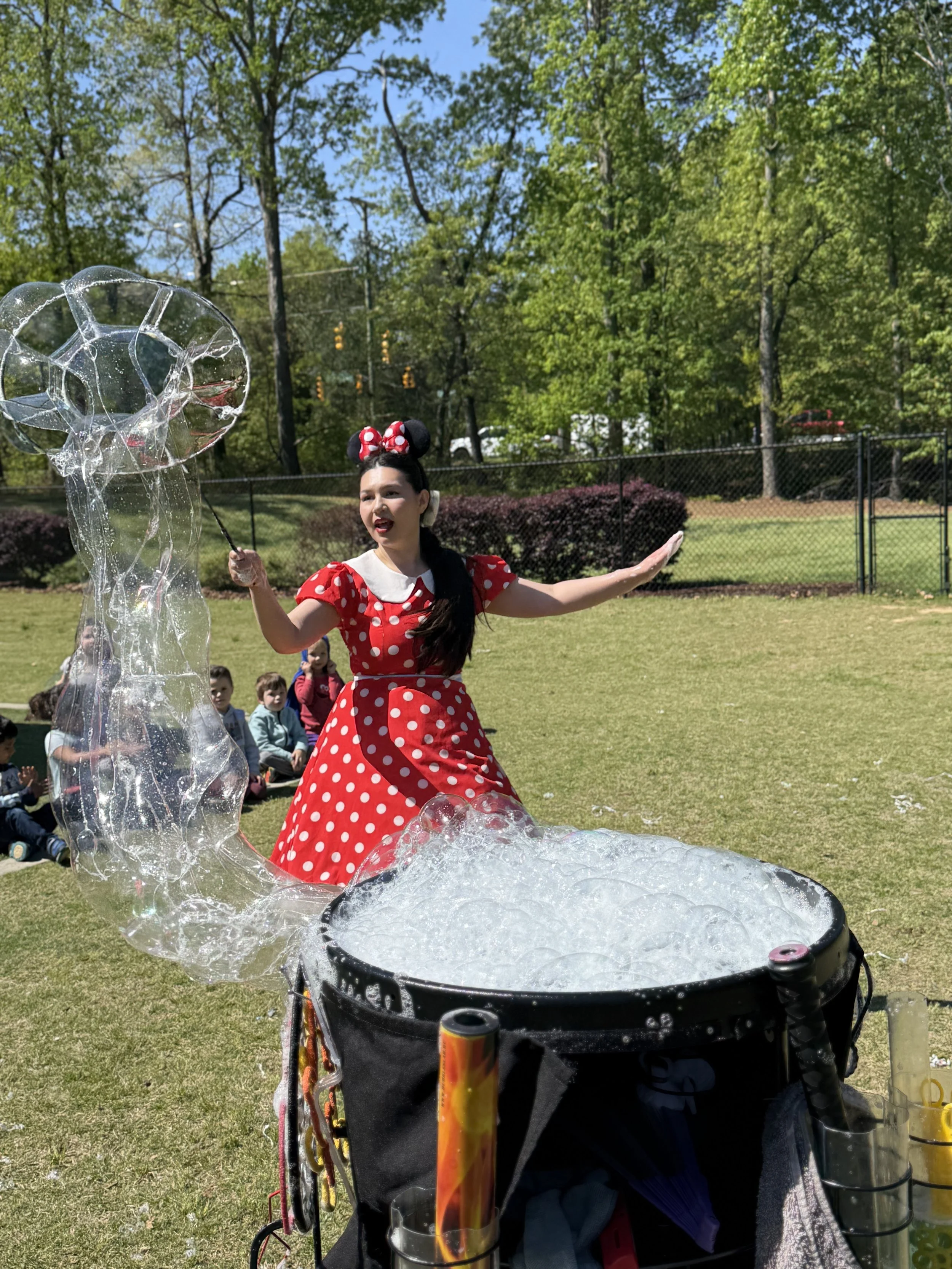 A woman dressed as Minnie Mouse making large soap bubbles outdoors in a park, with children sitting on the grass watching.