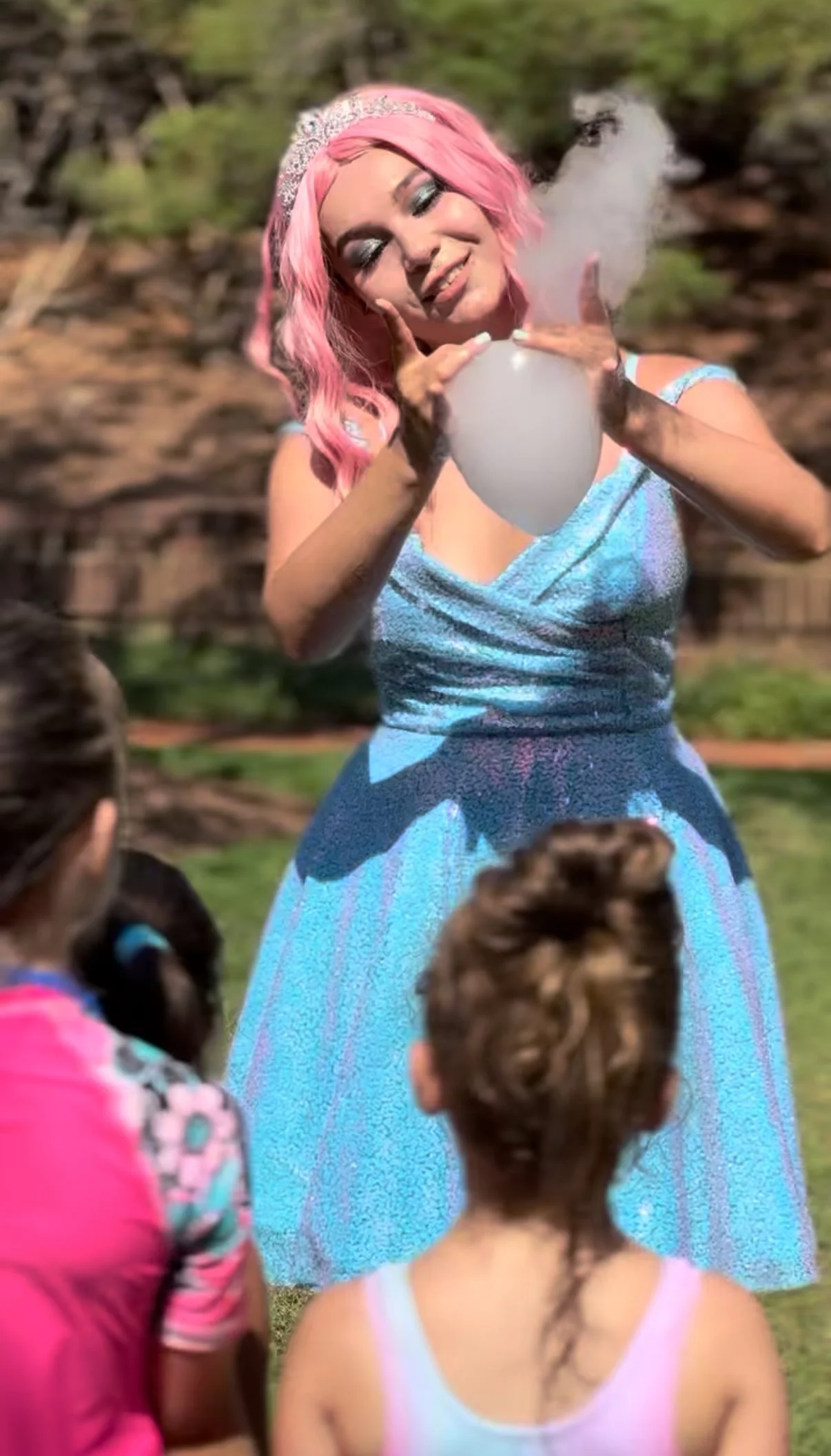 A woman with pink hair wearing a blue dress and a tiara blowing bubbles for children outdoors.