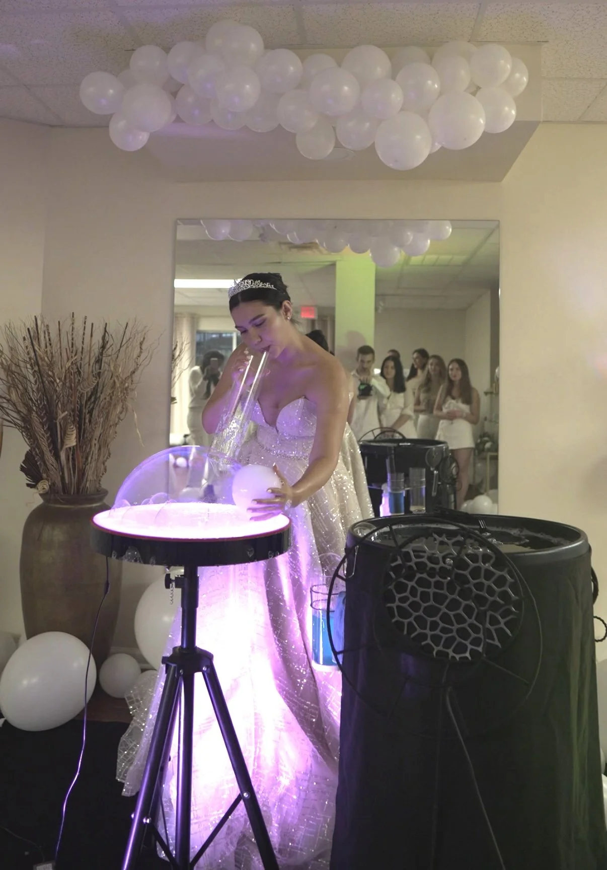 A woman in a sparkling dress wearing a tiara is performing a bubble show at a celebration, with white balloons and guests in the background.