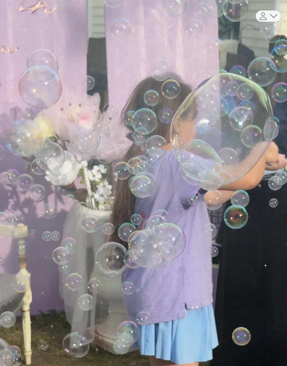 A girl with long hair and a purple top is playing with soap bubbles outdoors, with pink fabric and decorative flowers in the background.