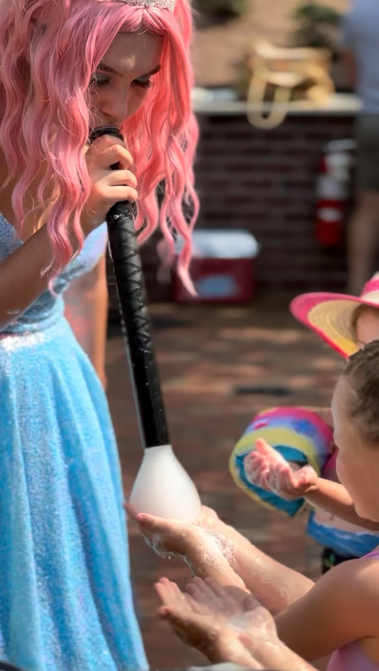 A girl with pink hair and a blue dress blows bubbles while children in swimsuits and a colorful hat wash their hands with soap and water.