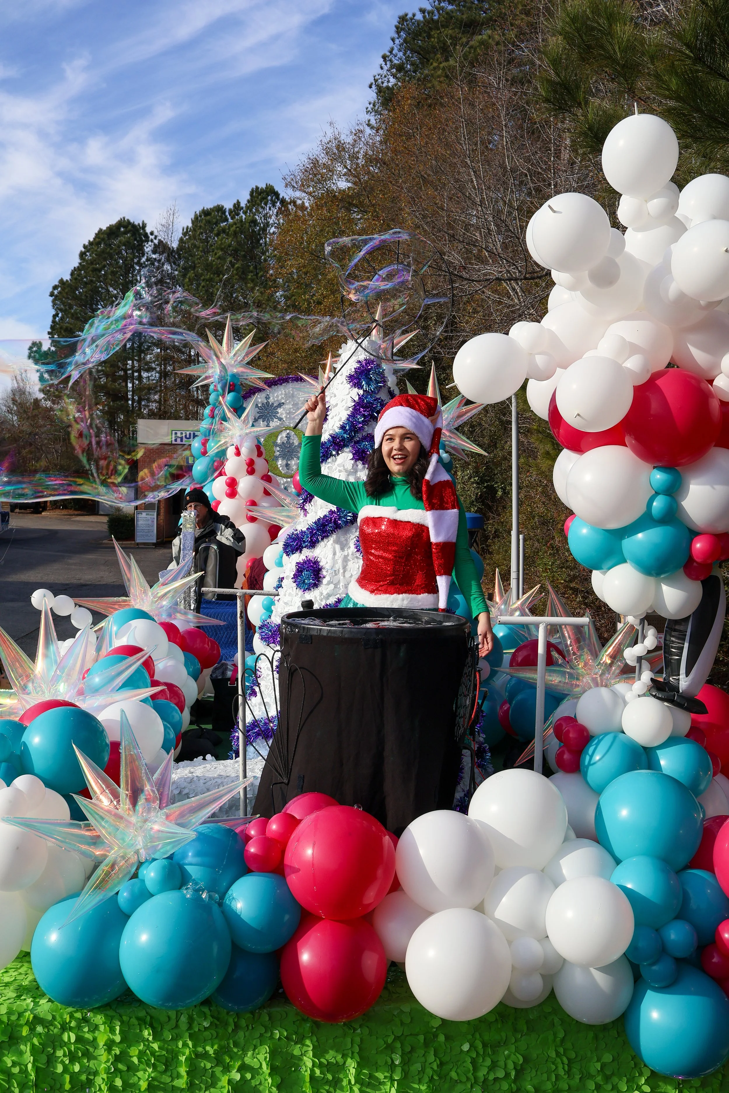 Woman wearing a Christmas hat and elf costume, smiling and waving from a decorated float with balloons and ornaments at a holiday parade.