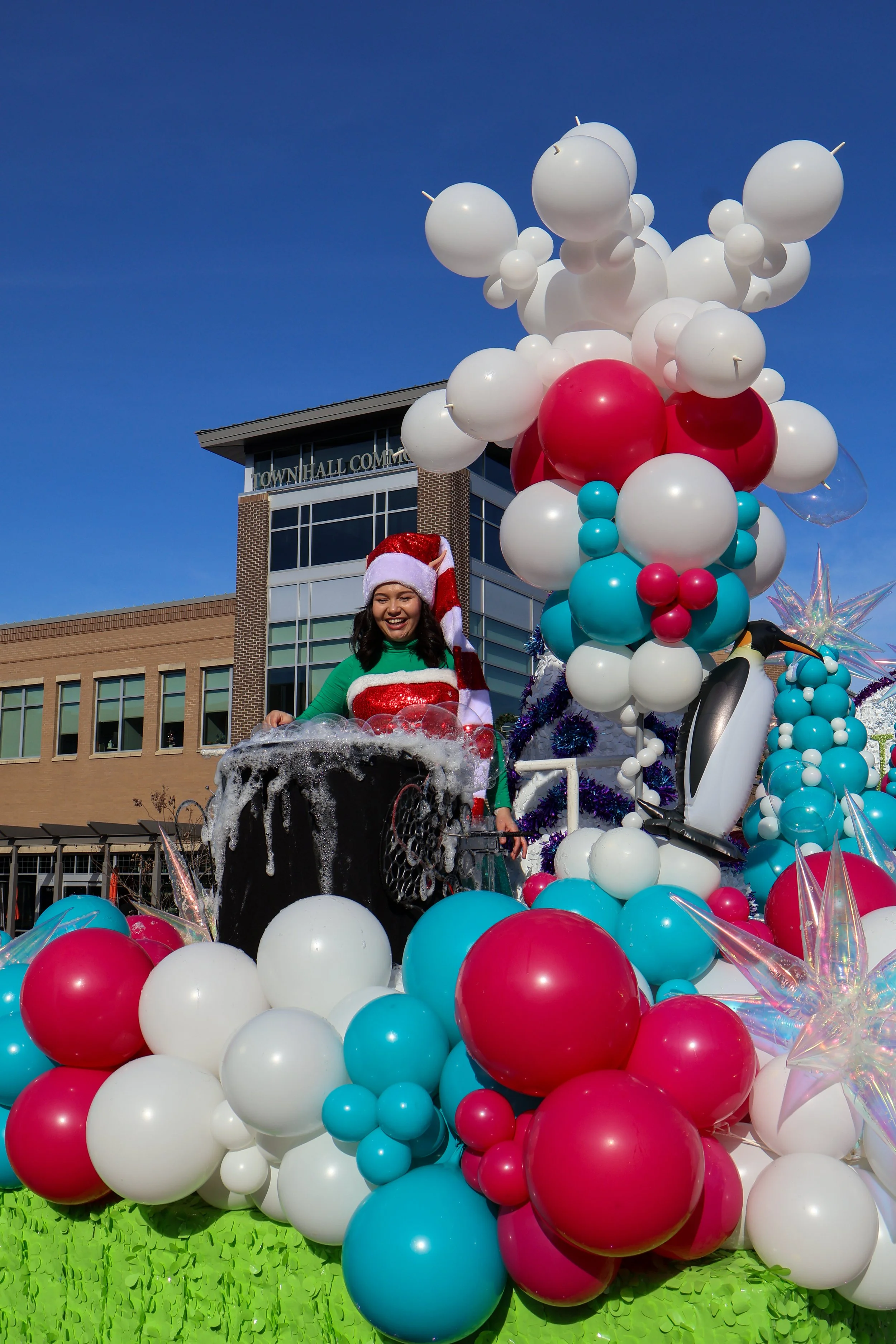 A woman in a Santa hat and festive sweater standing on a float decorated with colorful balloons, an inflatable penguin, and shiny ornaments during a parade.